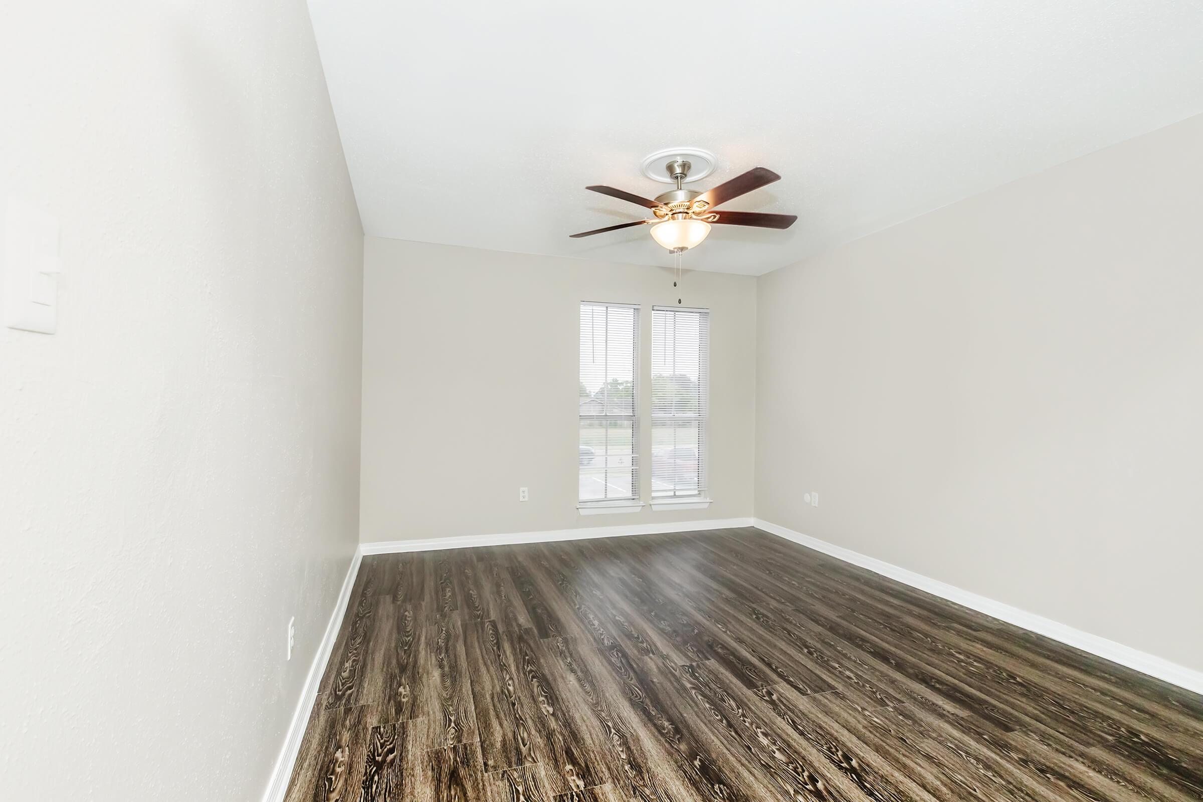 A spacious and unfurnished room with light-colored walls and a dark wood-like floor. There is a ceiling fan with light fixtures, and large windows with blinds allowing natural light to enter. The room has a clean and modern appearance, suitable for various uses.