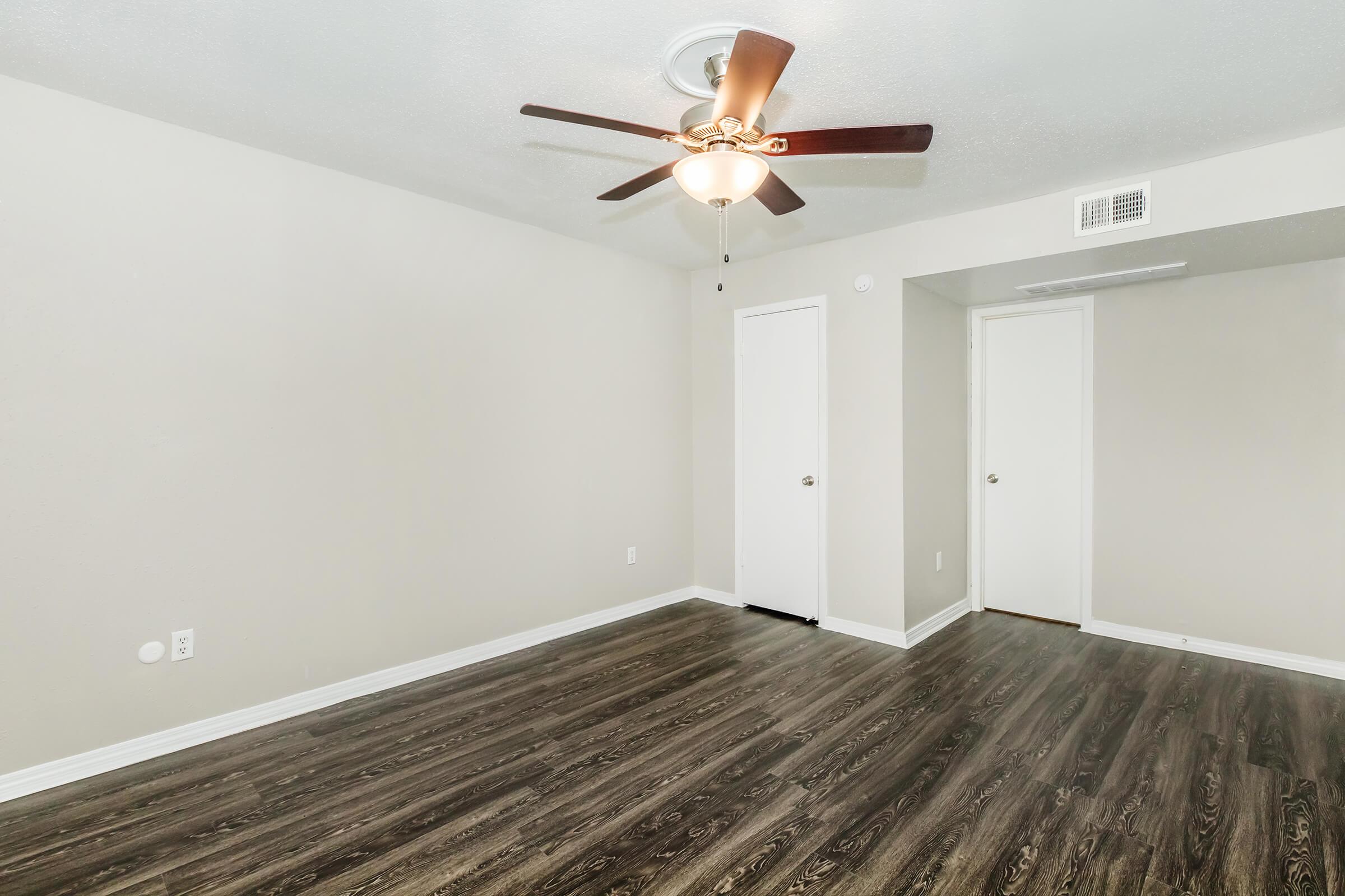 Interior of a light-colored room featuring a ceiling fan with wooden blades, wood-look laminate flooring, and white trim. There are two doors on the right, one leading to a closet, and the walls are painted a soft neutral tone, creating a spacious and inviting atmosphere.
