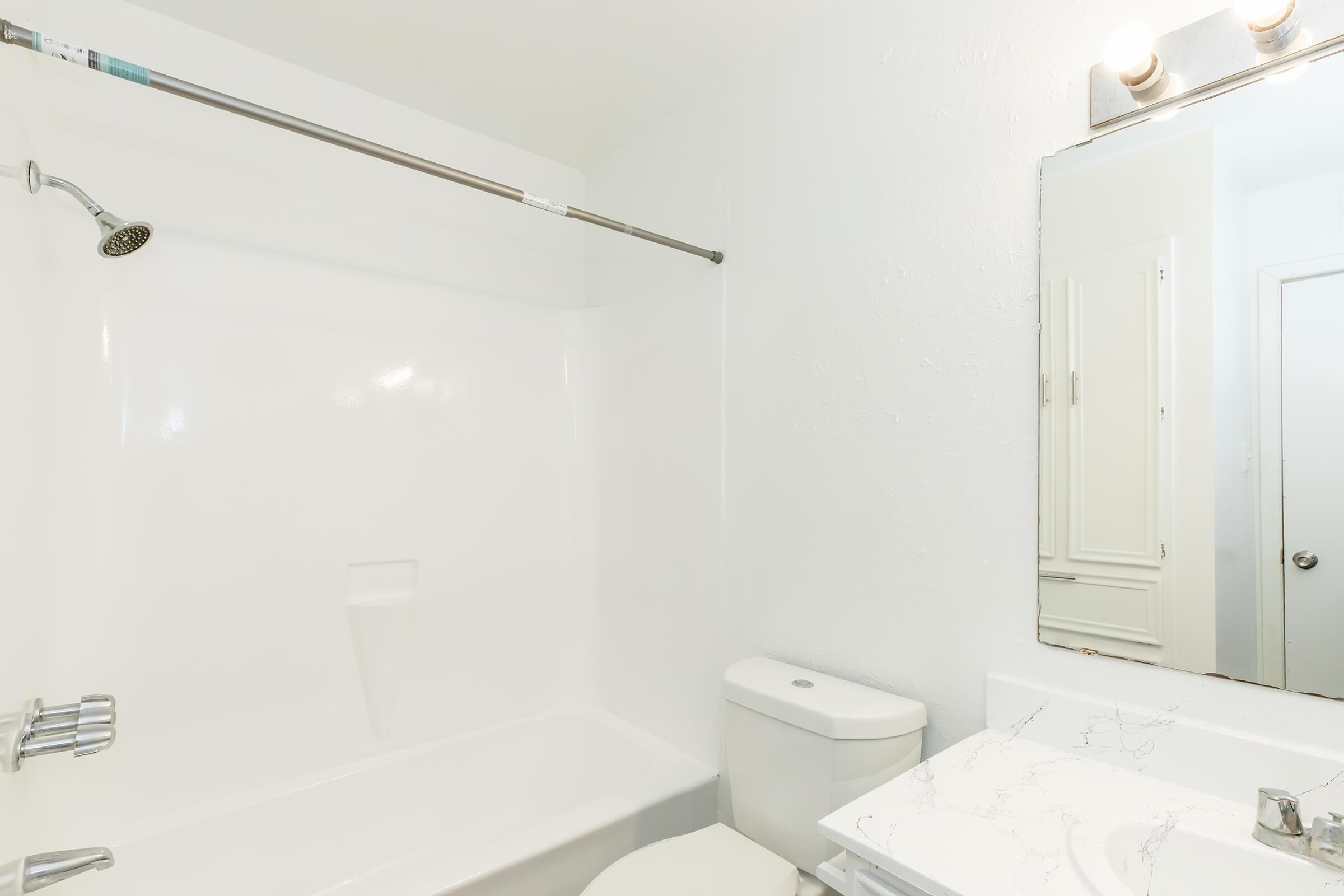 A clean, modern bathroom featuring a white bathtub with a showerhead, a white toilet, and a simple vanity with a marble-style countertop and mirror. The walls are painted white, and there is a light fixture above the mirror. The space is well-lit and uncluttered.