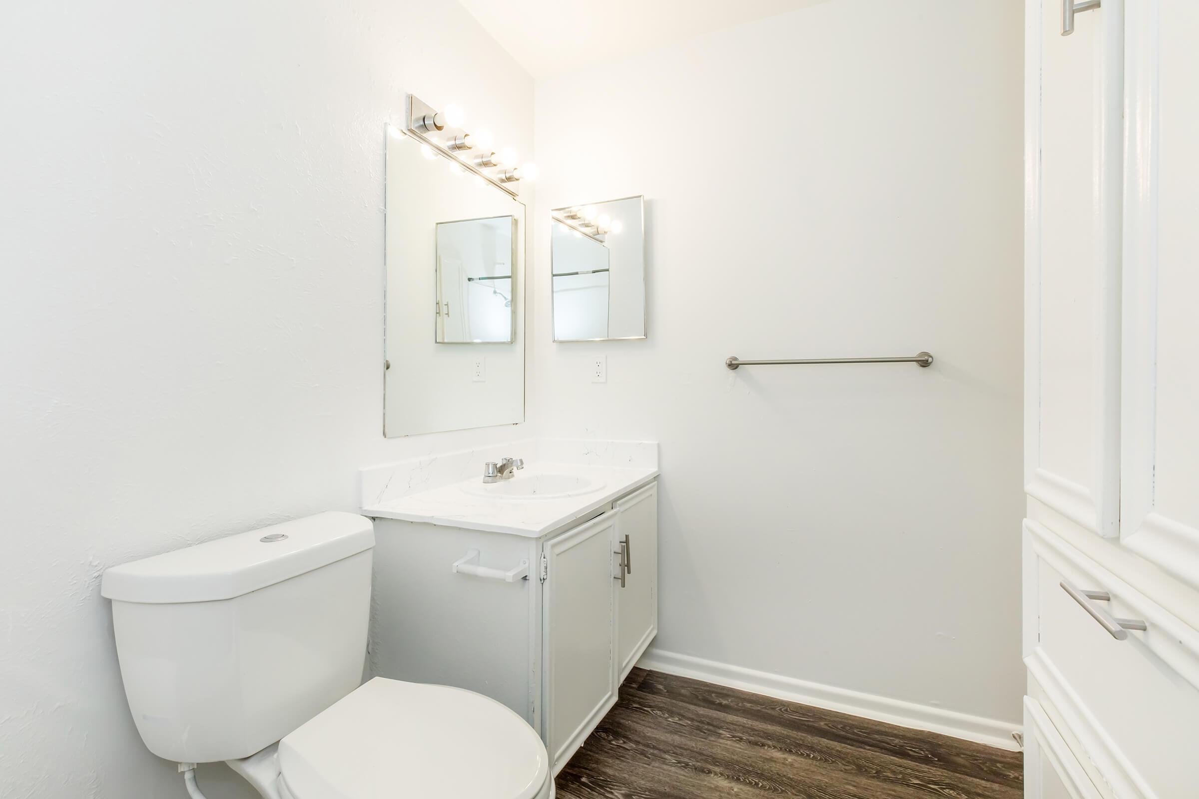 A clean and modern bathroom featuring a white toilet, a small vanity with a sink, a large mirror, and good lighting. The walls are painted in a light color, and the floor has a dark wood finish, creating a bright and spacious atmosphere.