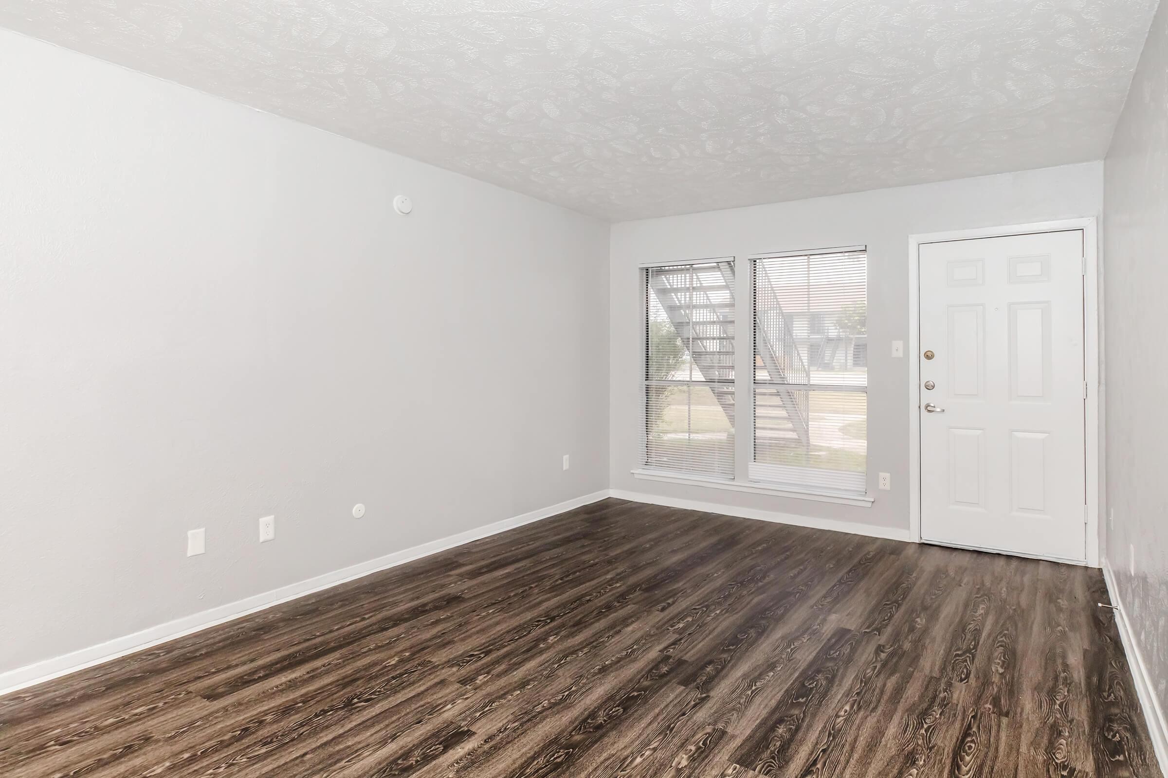 An empty living room featuring light gray walls, a textured ceiling, large windows with blinds, and a wooden floor. There is a front door on the right, and the room is well-lit with natural light coming through the windows.