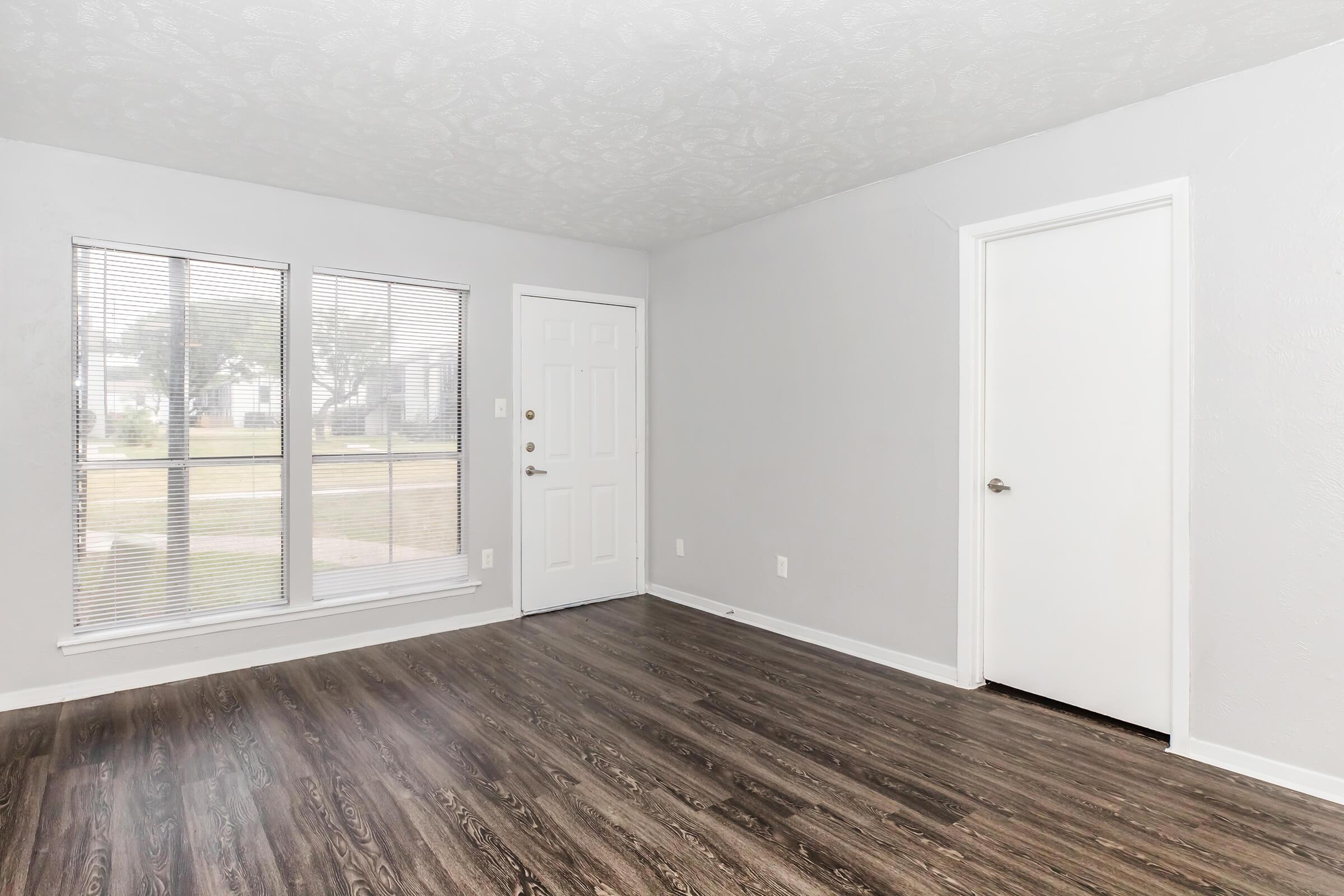 Empty living room with light gray walls, vinyl flooring, and large windows allowing natural light. A front door is visible, along with a closet door on the right. The space is clean and uncluttered, creating a bright and inviting atmosphere.