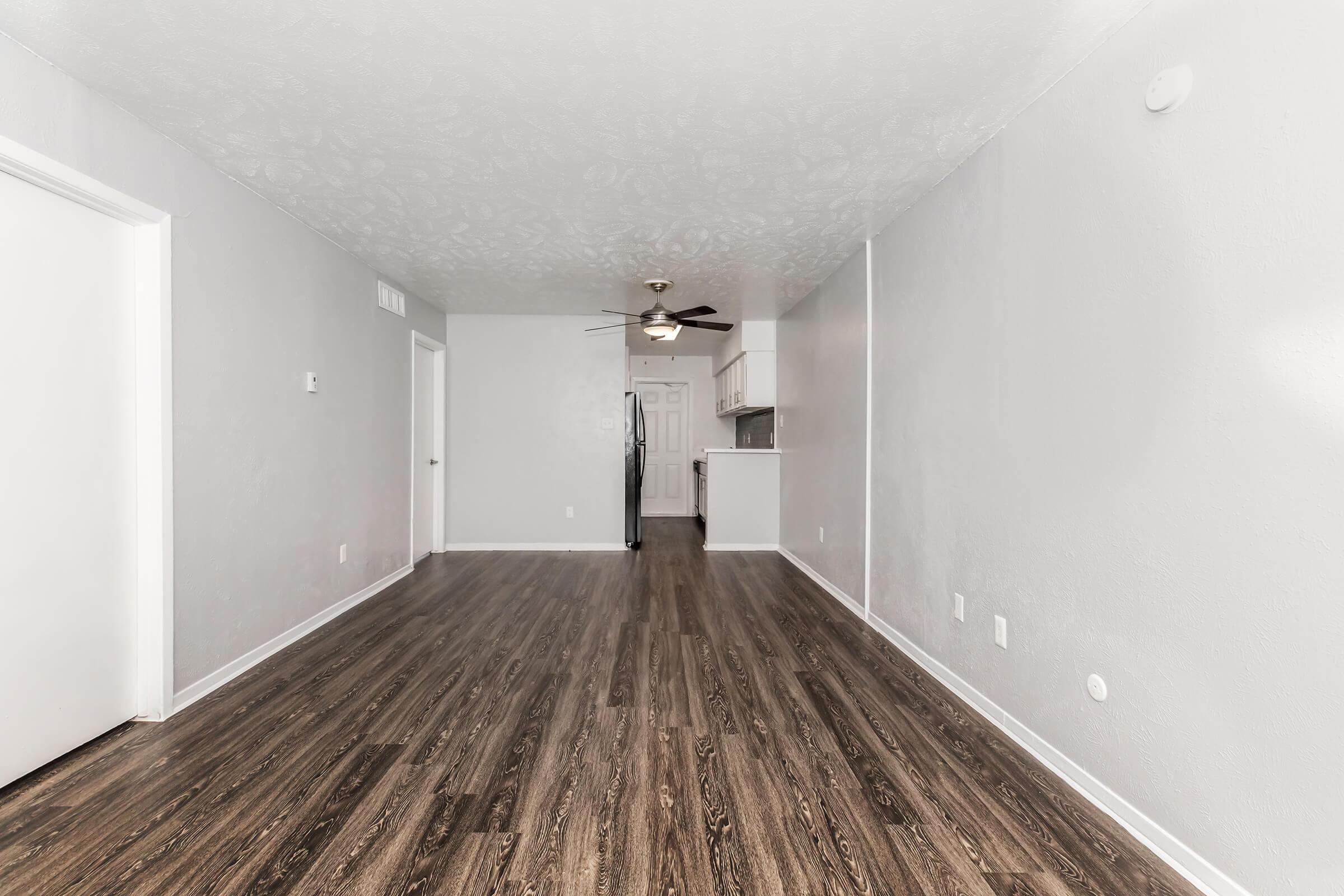 A spacious, empty living room with gray walls and dark wood-like flooring. A ceiling fan is visible, and a kitchen can be seen in the background. The room is well-lit, creating an open and airy atmosphere. There are two doors on the left side, suggesting access to other spaces.