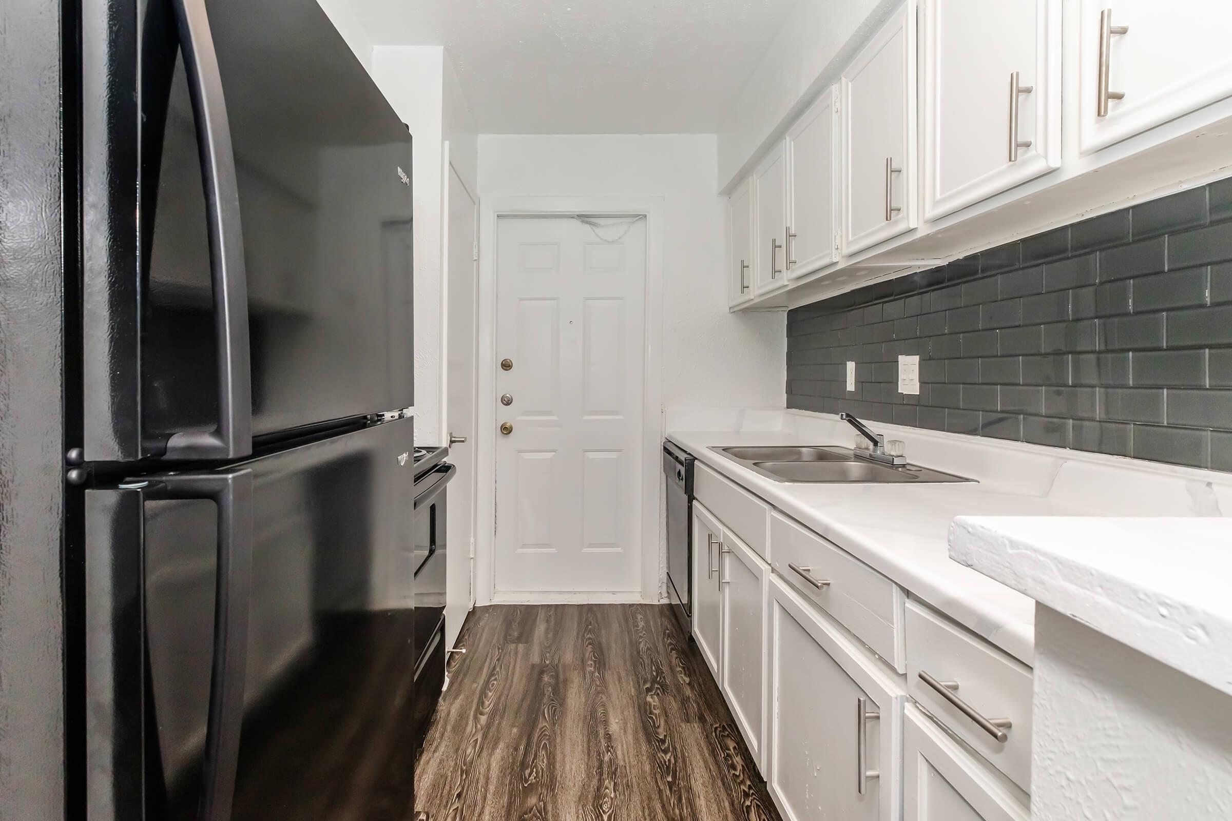 A modern kitchen featuring white cabinetry, a black refrigerator, and stainless steel appliances. The countertop is white, and there is a dark tiled backsplash. The room includes a door leading to an exterior space and has laminate flooring in a wood design. Natural light illuminates the area.