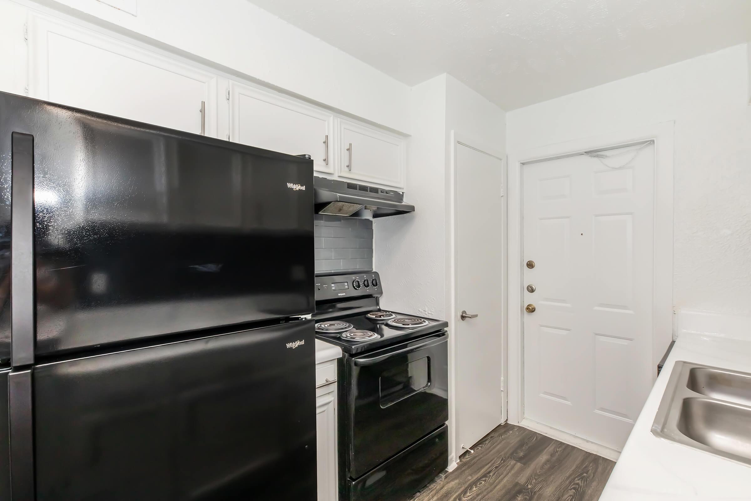 A kitchen featuring a black refrigerator, a black stove, and a stainless steel sink. The cabinetry is white, providing a modern contrast to the appliances. A door leads to another area, and the flooring appears dark, enhancing the sleek design of the space.