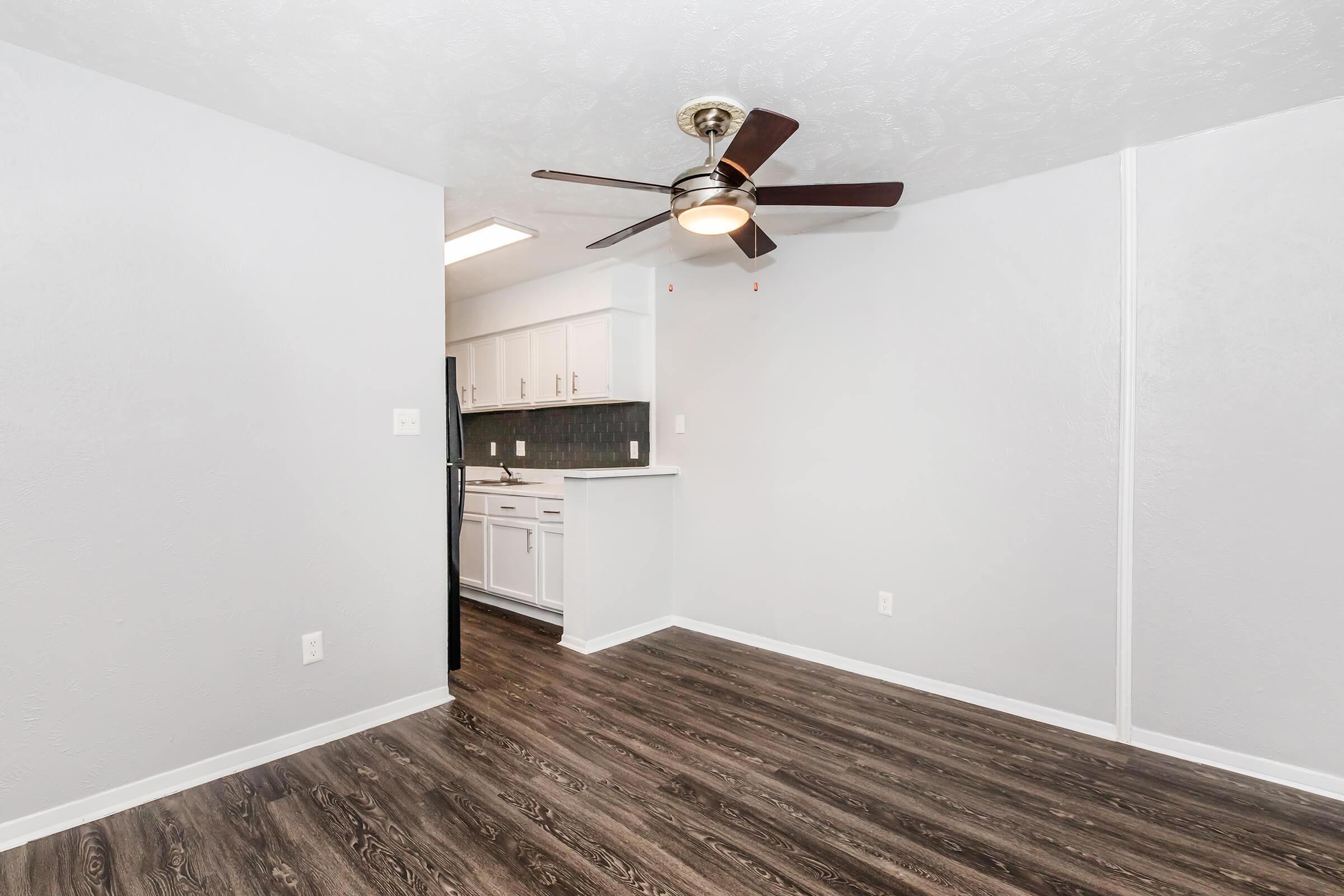 A modern living space featuring a light gray wall color, dark wood-style flooring, a ceiling fan, and a compact kitchen area with white cabinets and a dark backsplash in the background. Natural light comes from a ceiling fixture, creating a bright and inviting atmosphere.