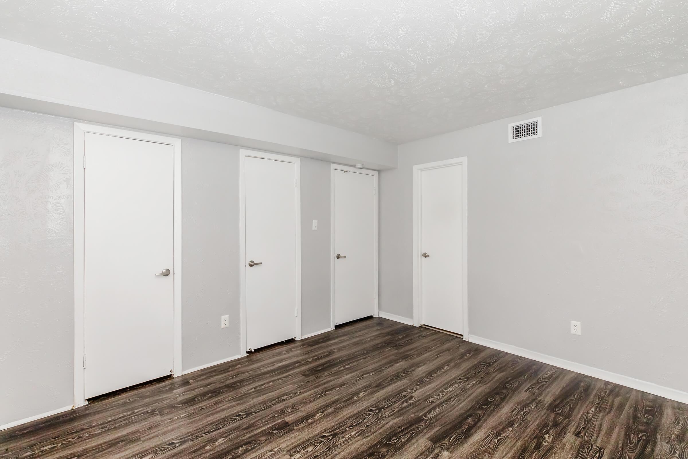 A spacious, empty room with light gray walls and a textured ceiling. There are four white doors lined up against one wall, and the floor features dark wood laminate. The overall atmosphere is clean and neutral, suitable for various uses.