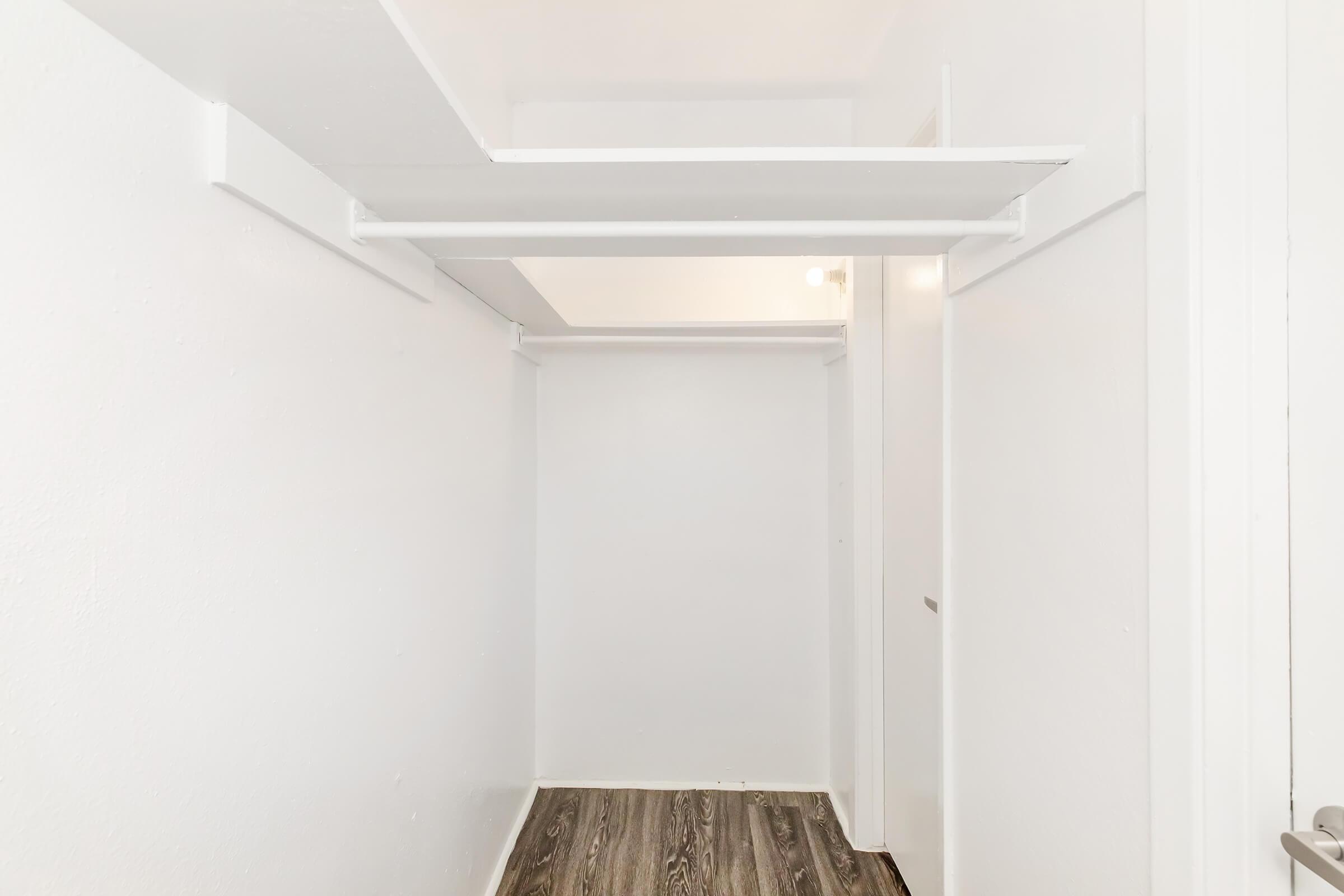 Empty interior space of a closet with white walls and a wooden floor. The closet features overhead shelving and a small opening leading to another room. Natural light is visible, creating a bright and clean atmosphere.