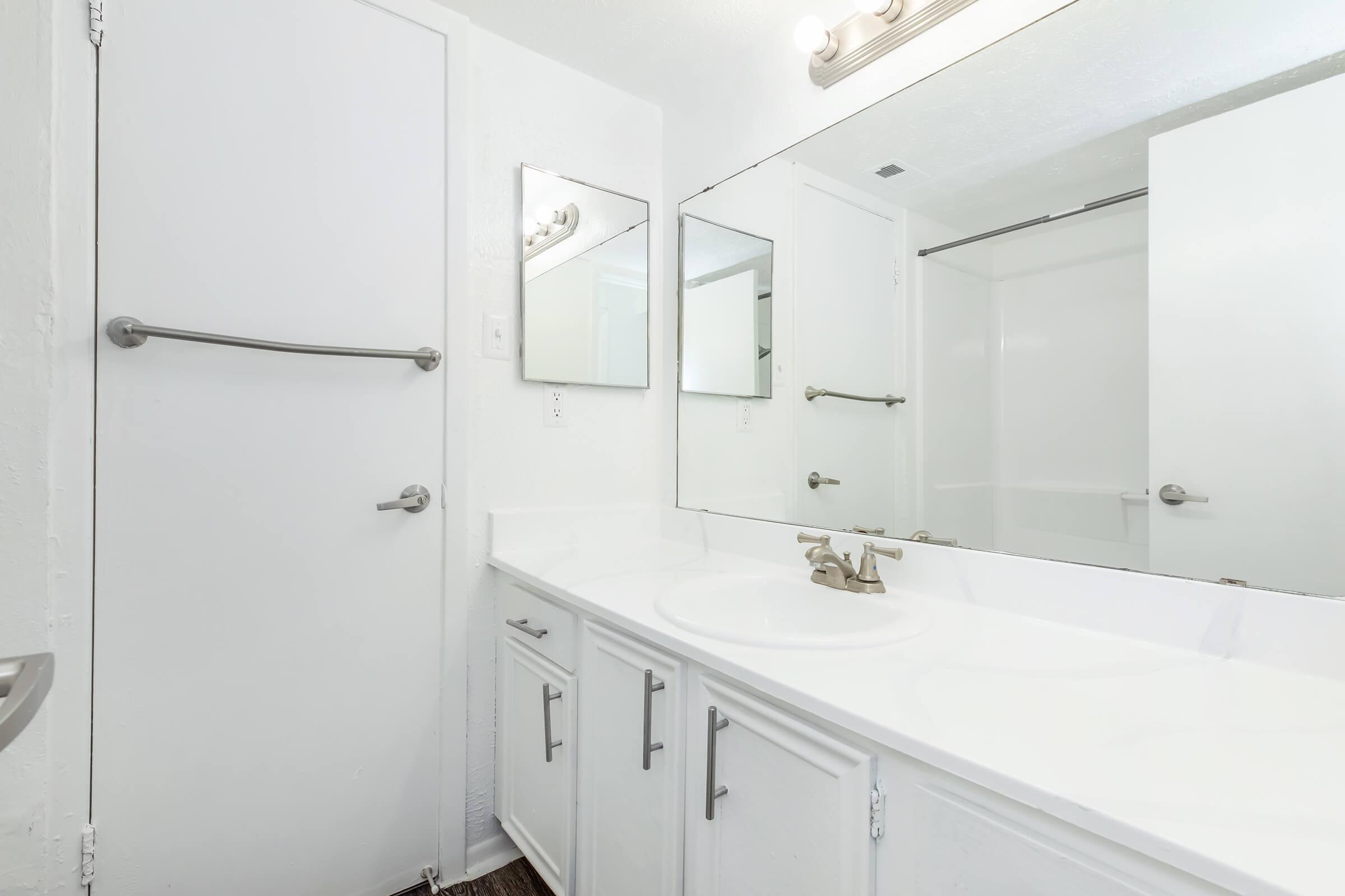 A clean, well-lit bathroom featuring a white countertop with a sink, two mirrors above, and a sliding shower door. The walls are painted white, and there is a towel bar mounted on the wall. The overall appearance is modern and minimalistic.