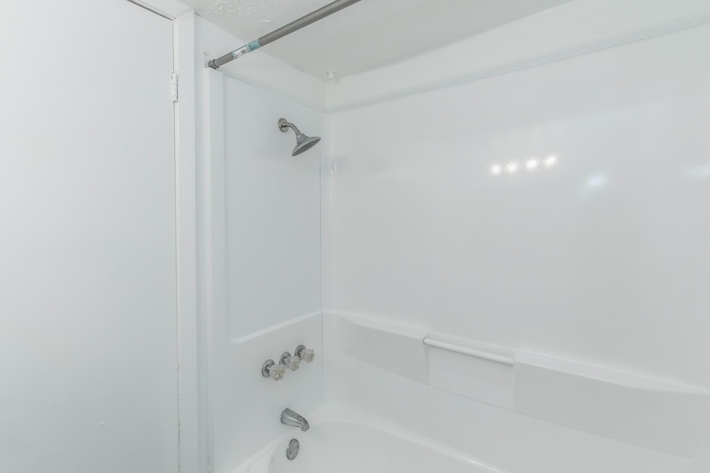 A clean, minimalist bathroom featuring a shower area with a wall-mounted showerhead, a built-in bathtub, and smooth white walls. The door to the bathroom is partially visible, and there are no visible toiletries or decorations. The lighting is bright, creating an airy atmosphere.
