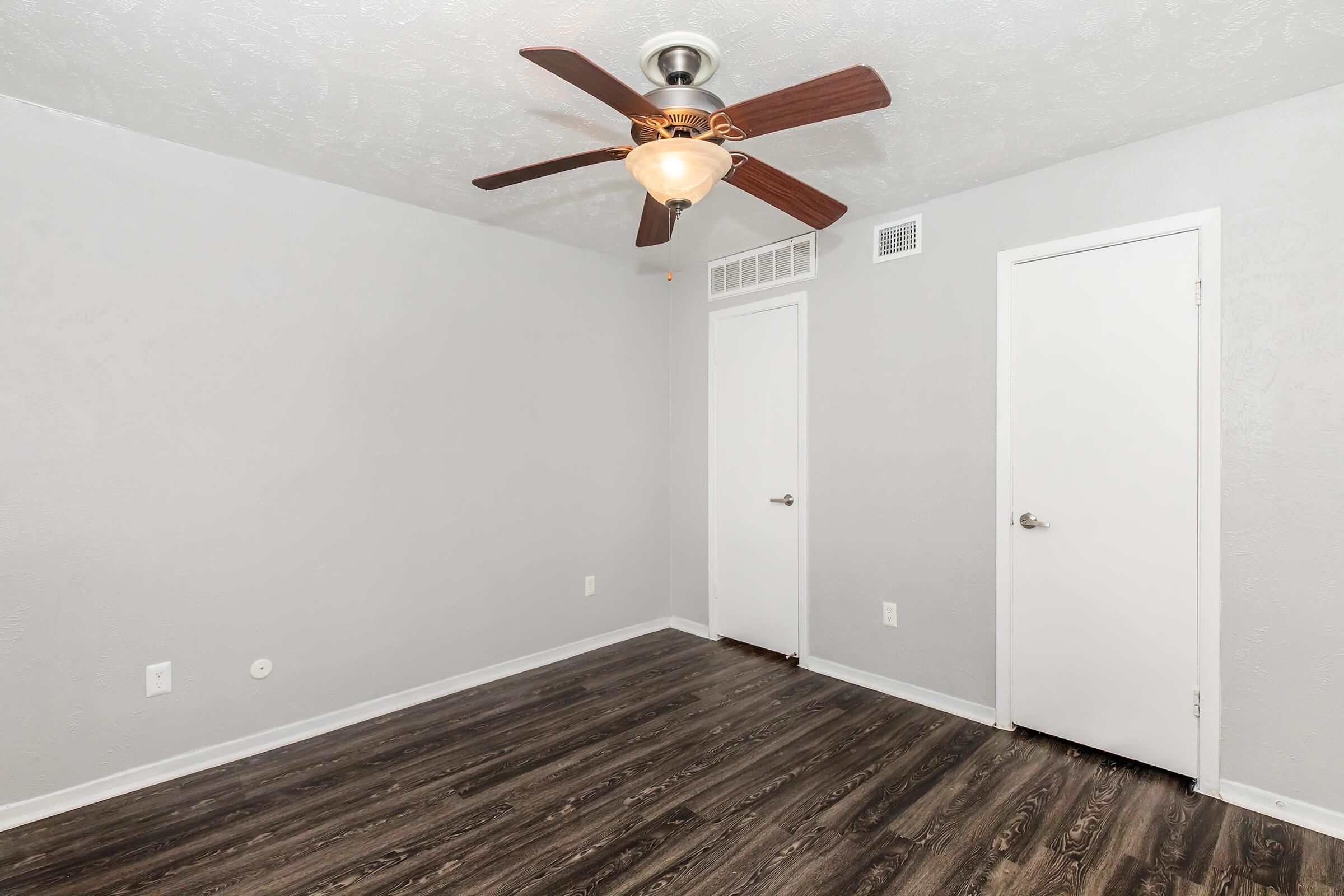 An empty room with gray walls and dark wood-like flooring. It features a ceiling fan with a light fixture and has two white doors on the right side. The space is well-lit and has a clean, modern appearance.