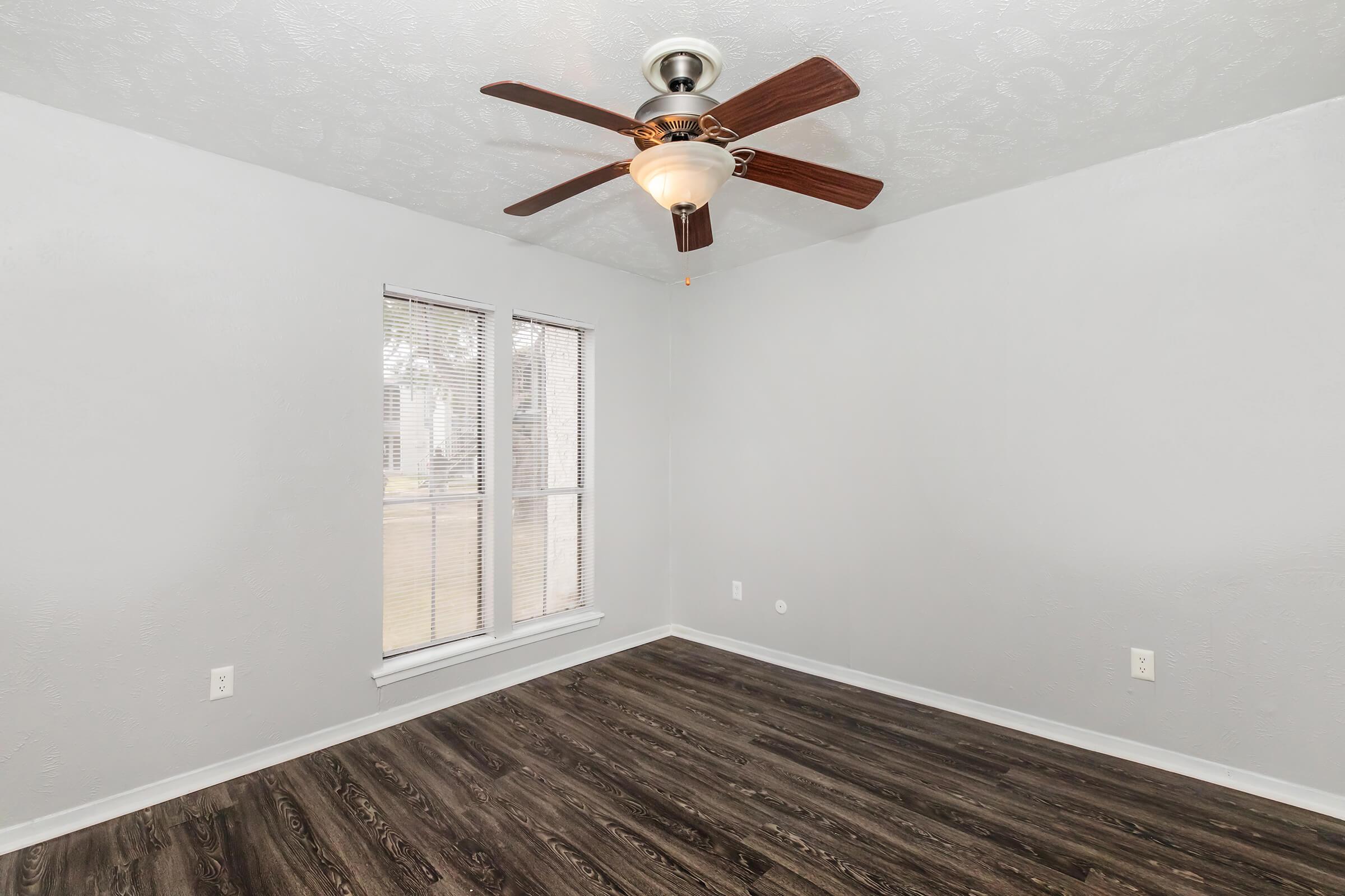 Empty room featuring light gray walls and dark wood-style flooring. A ceiling fan with a light fixture is centered in the room, and there are two windows on one wall allowing natural light to enter. The space has a clean and modern appearance, making it suitable for various uses.