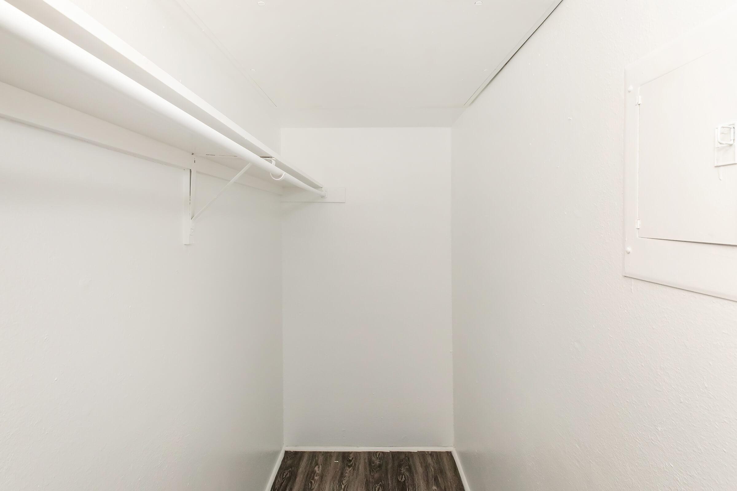 A small, empty closet with white walls, a single white hanging rod, and a light-colored floor. There is a small panel on one wall, and the overall space appears minimalist and unoccupied.