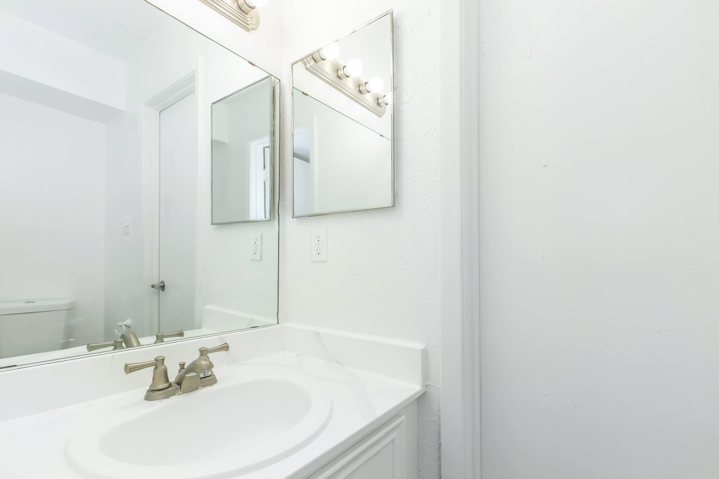 A clean, bright bathroom featuring a white countertop with a sink, dual mirrors above the sink, and a light fixture. The walls are painted white, and there's a glimpse of a door and a toilet in the background, creating a serene and uncluttered atmosphere.