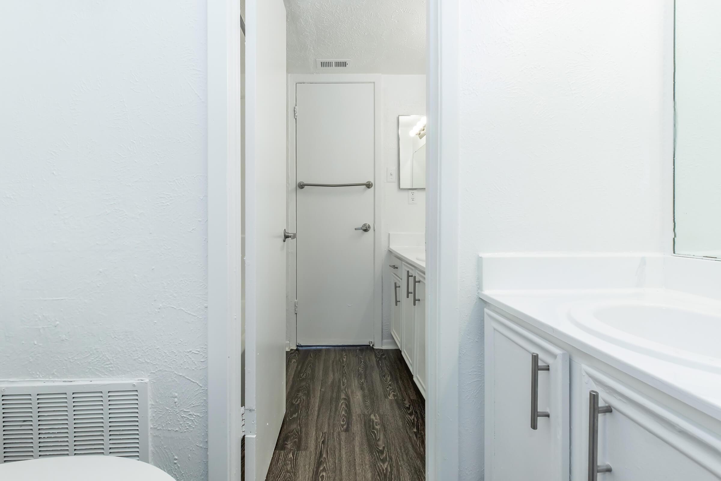 A clean, modern bathroom featuring white walls, a white vanity with a sink, and a mirror. The hallway leads to a closed door at the end, with dark wood-like flooring throughout. Bright lighting enhances the spacious feel of the area.