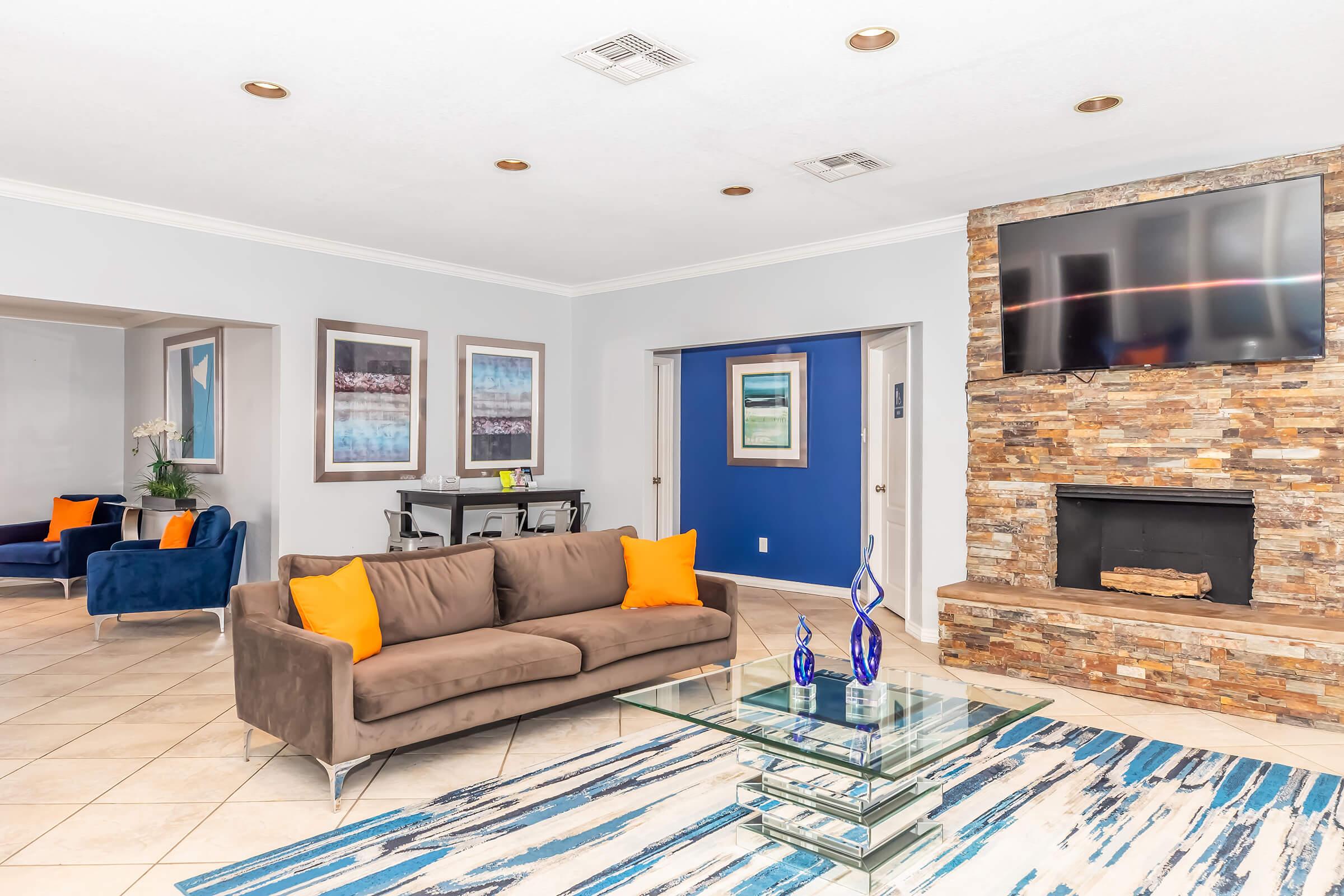 Modern living room featuring a brown sofa with orange cushions, a glass coffee table, and a decorative area rug in blue and white. There’s a stone fireplace with a mounted TV above it, framed artwork on the walls, and two blue chairs in the background. Natural light brightens the space.