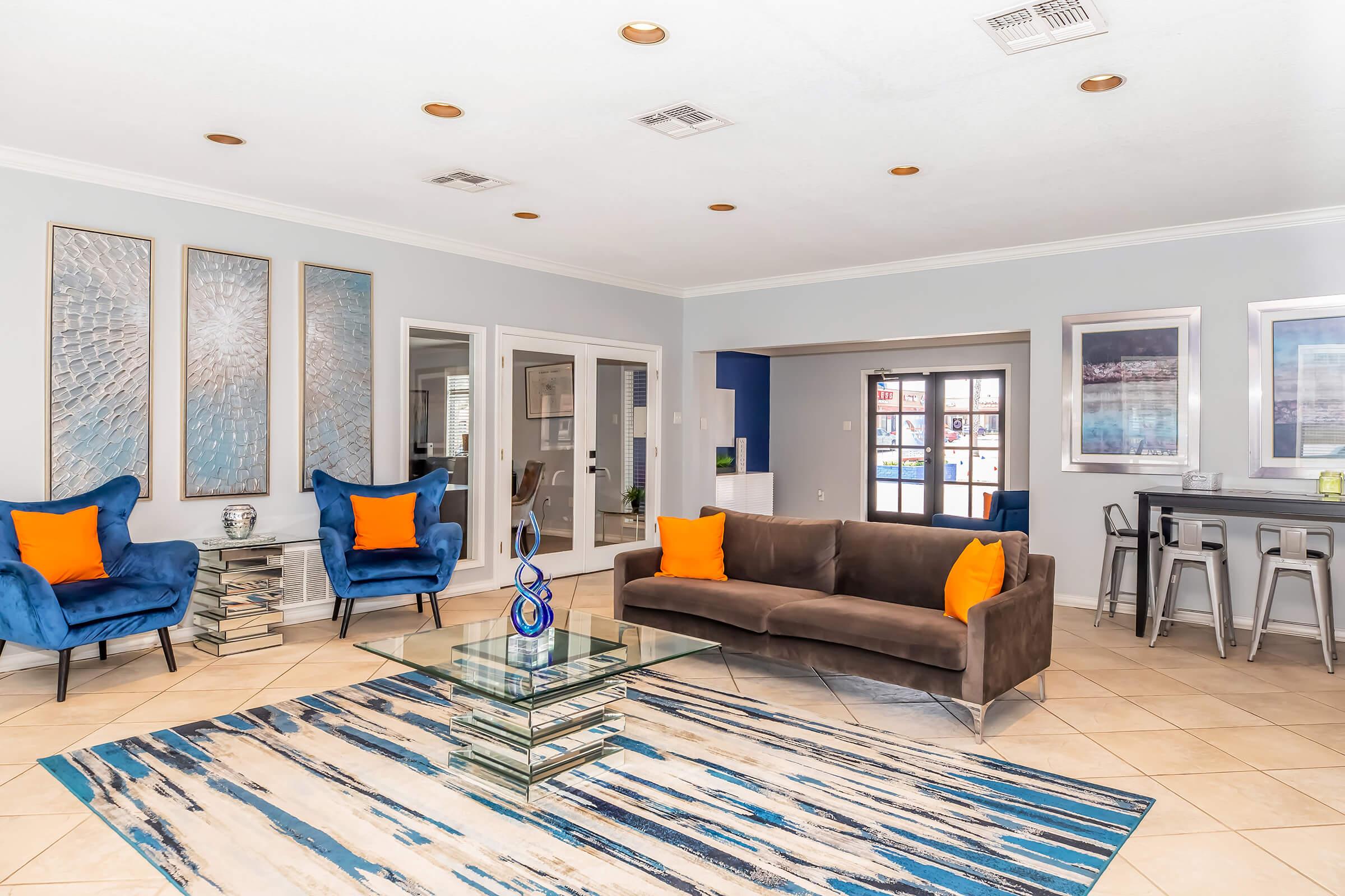 Modern living room featuring two blue chairs with orange cushions, a brown sofa, and a glass-top coffee table on a patterned rug. The room has light gray walls, decorative wall art, and large windows providing natural light. A sleek dining area is visible in the background.