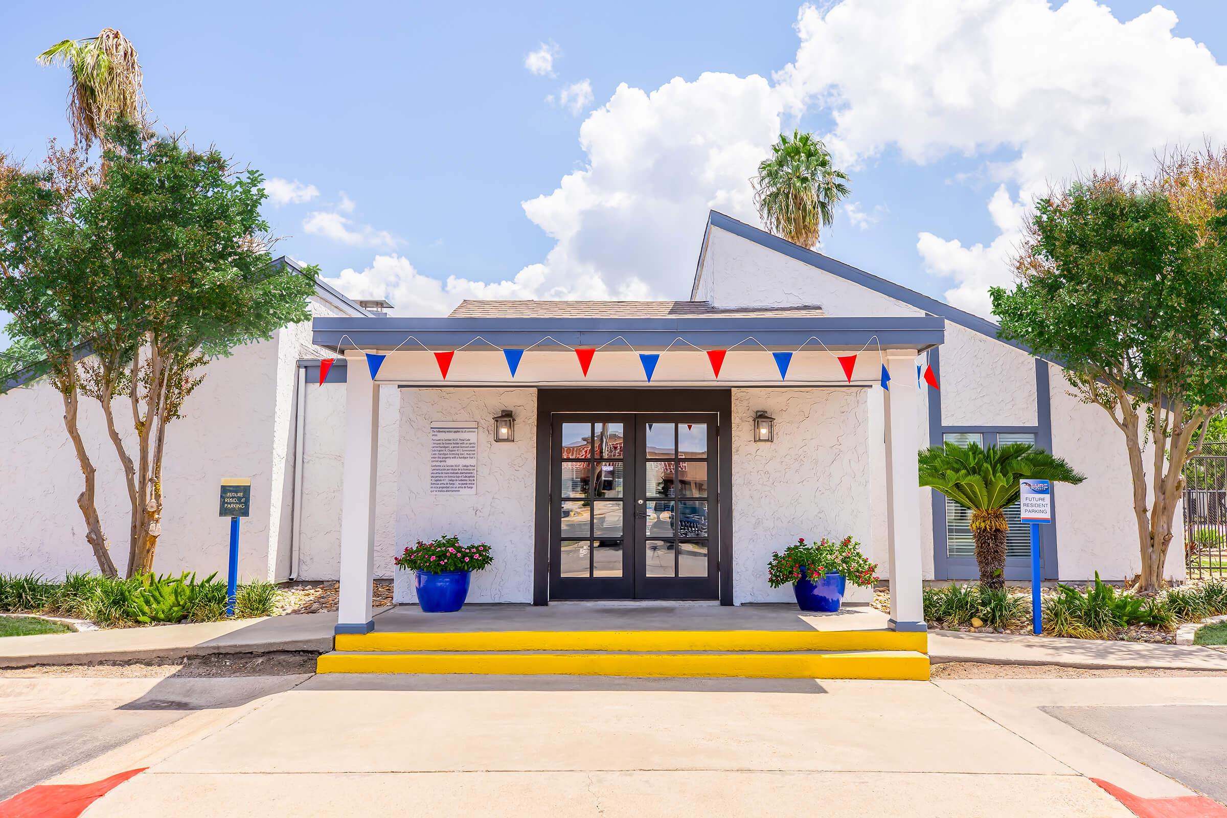 A white building entrance with a flat roof, featuring large glass doors framed by decorative lighting. The entrance is adorned with red, white, and blue bunting, and there are two blue pots with plants on either side of the door. Palm trees and greenery are visible in the surrounding area under a bright blue sky with fluffy clouds.