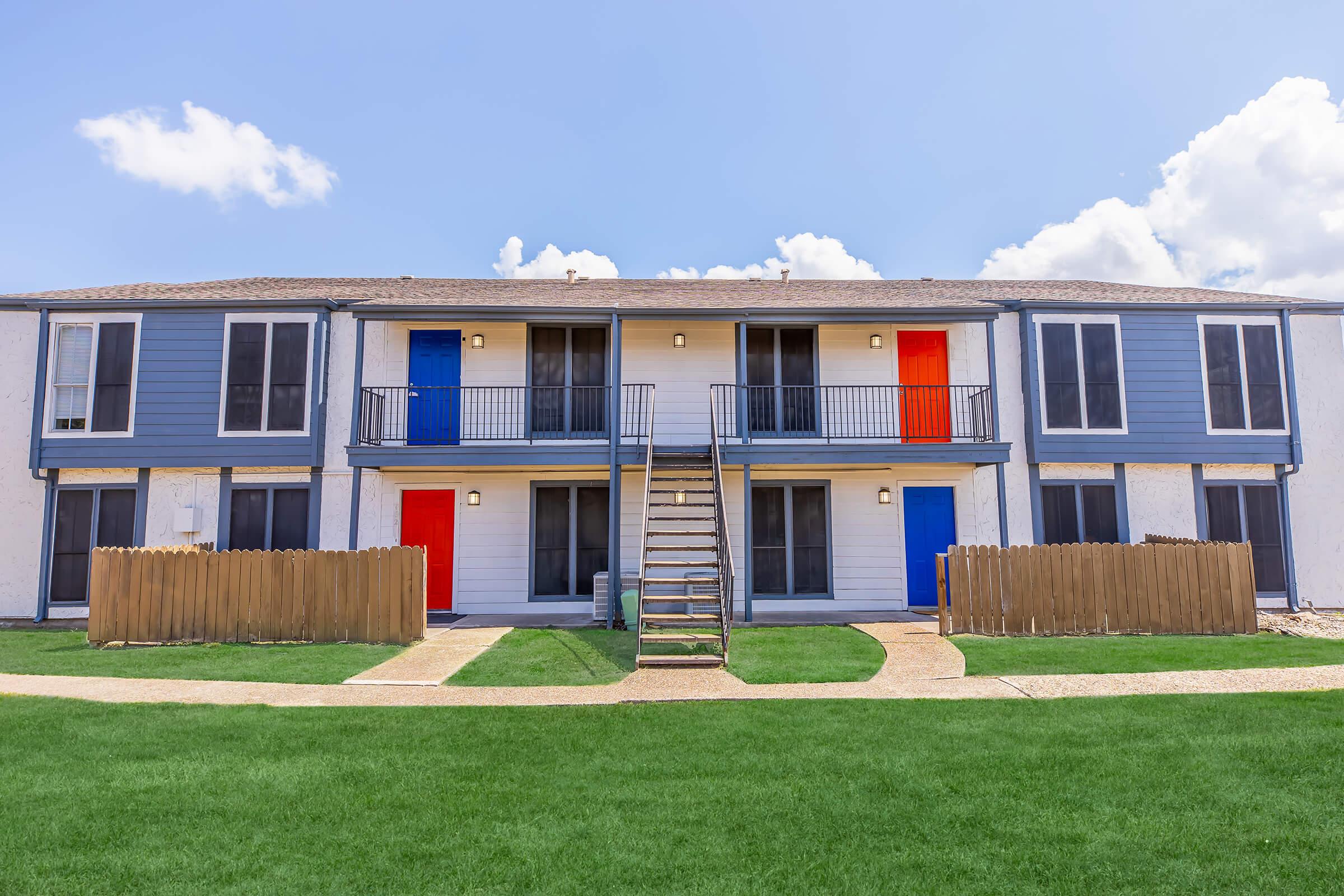Two-story apartment building with blue and orange doors, set against a clear blue sky. The building features a central staircase leading to the upper level, with well-maintained green grass in the foreground and wooden fencing surrounding the property.