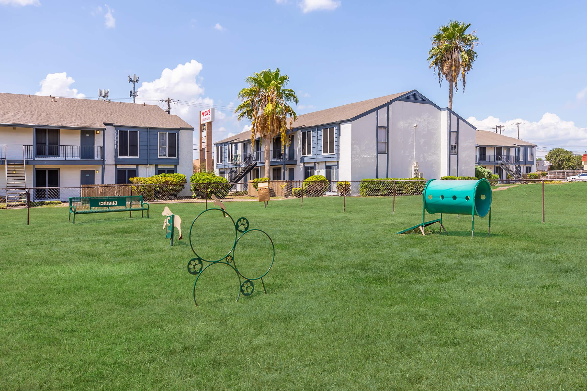 A grassy area featuring various dog agility equipment, including a tunnel, circular jump, and a small bench. In the background, there are two multi-story apartment buildings with palm trees and clear blue skies.
