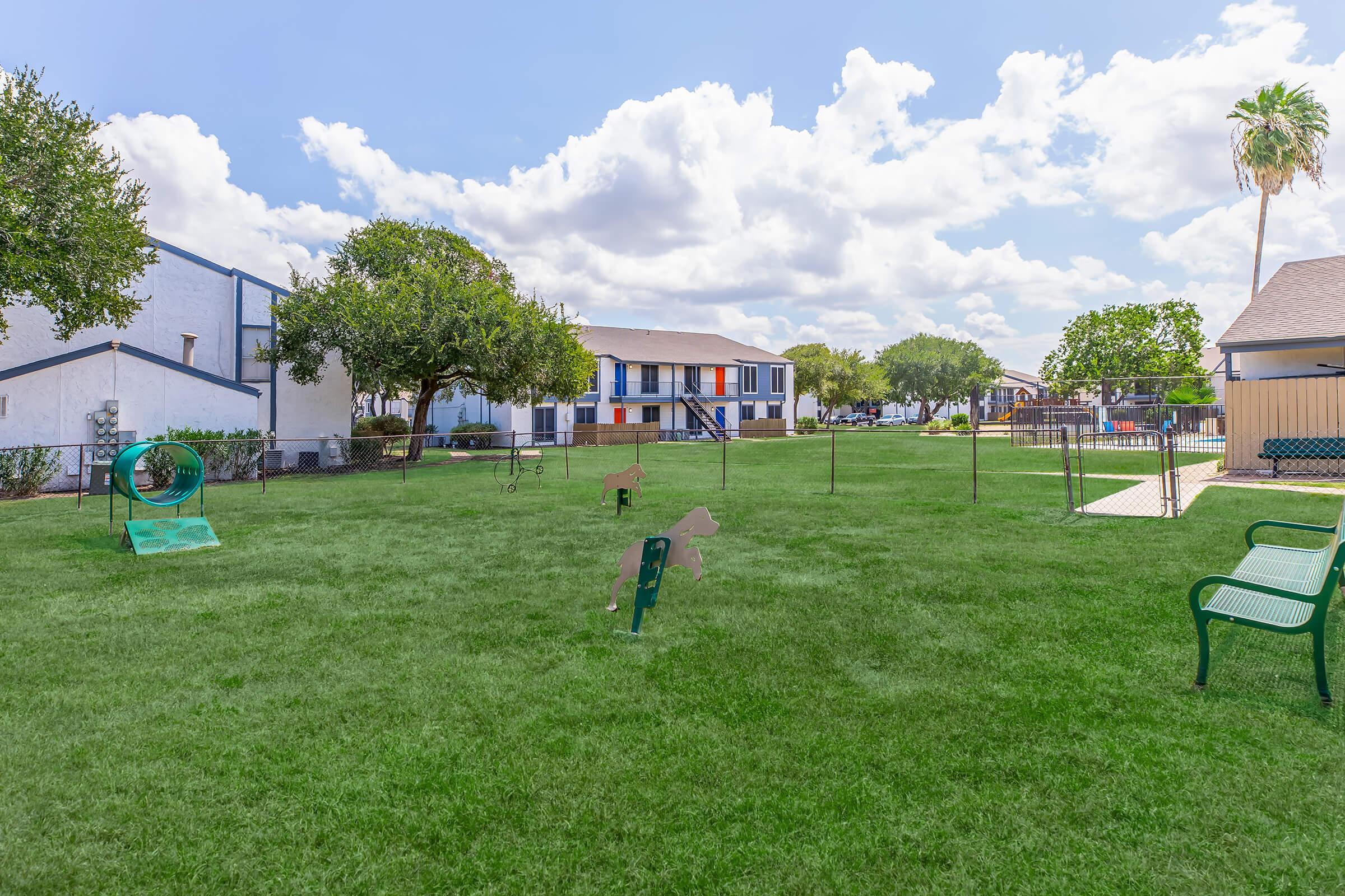 A grassy park area with benches, playful dog structures, and a fence, surrounded by residential buildings. The sky is partly cloudy, creating a bright, inviting atmosphere. Trees are present, enhancing the outdoor environment.