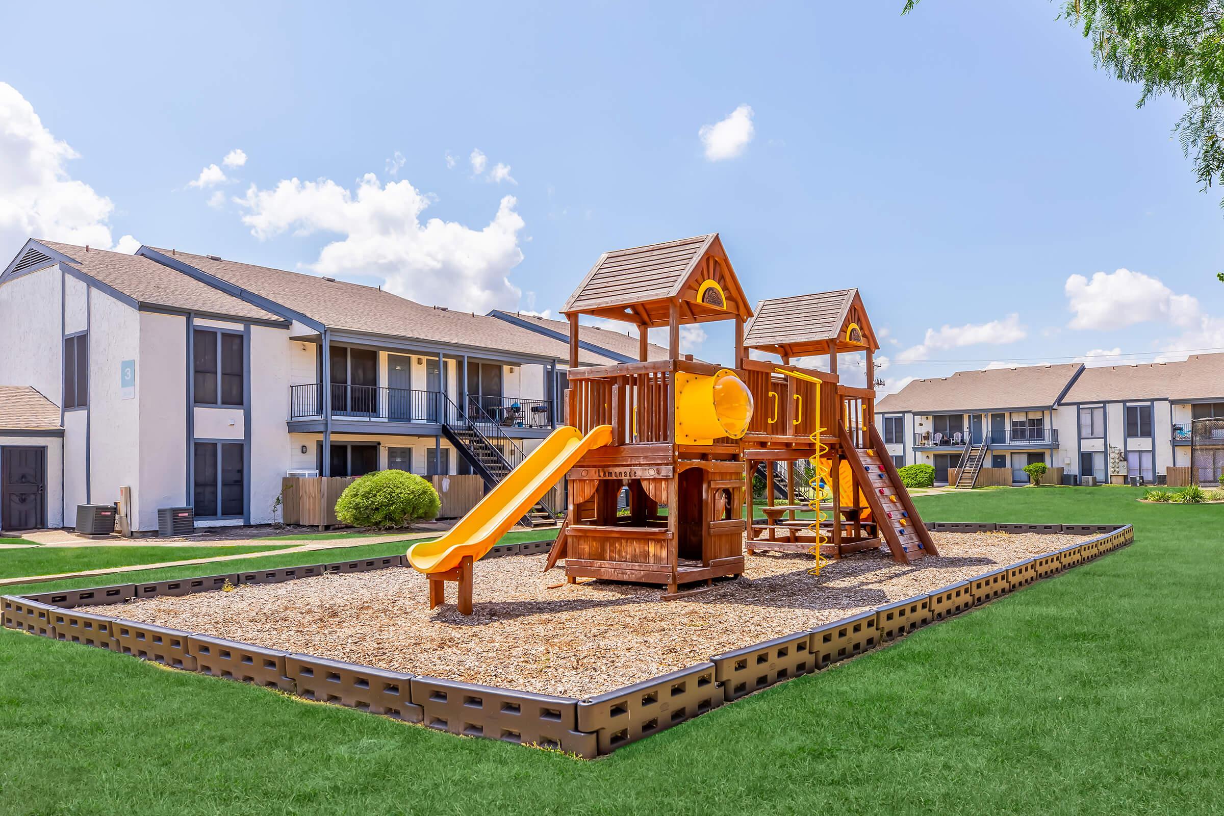 A wooden playground with a slide and climbing structure on a grassy area, surrounded by residential buildings. The sky is clear with fluffy clouds, and the scene conveys a bright, sunny day suitable for outdoor play.