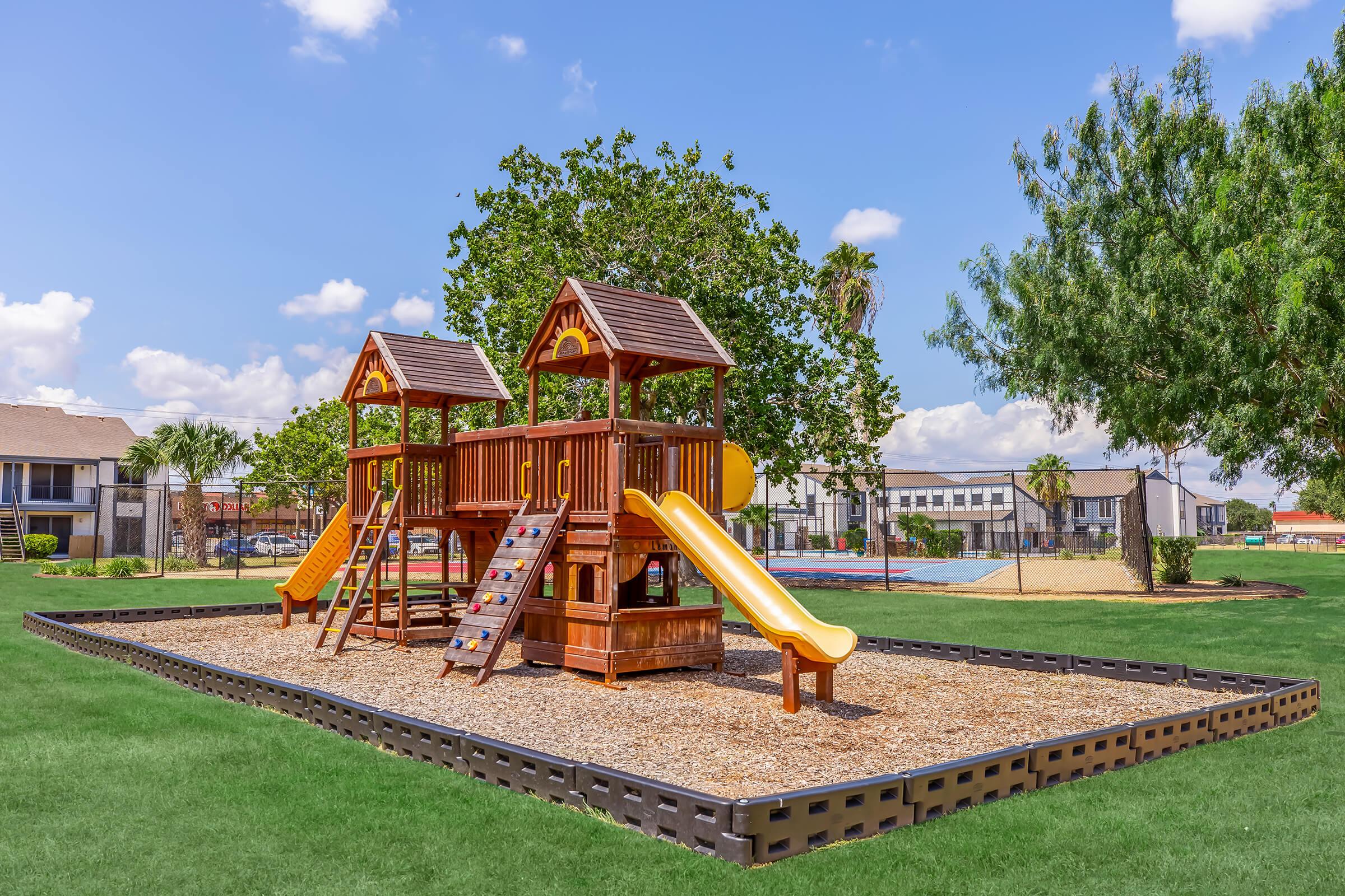 A wooden playground structure featuring two slides, ladders, and climbing areas, set on a grassy field. The background includes trees, a sandy play area, and a building, with a clear blue sky and fluffy clouds above.