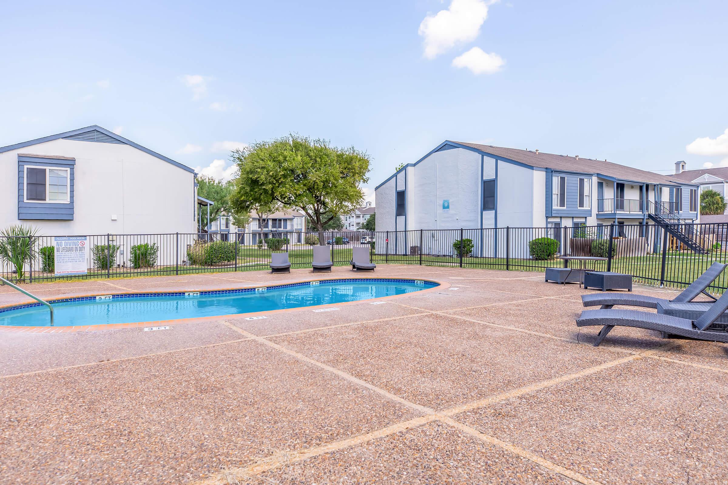 A clear swimming pool surrounded by a patio area with lounge chairs. Two multi-story residential buildings are visible in the background, along with green grass and trees. The scene is bright and sunny, creating a relaxed outdoor atmosphere.