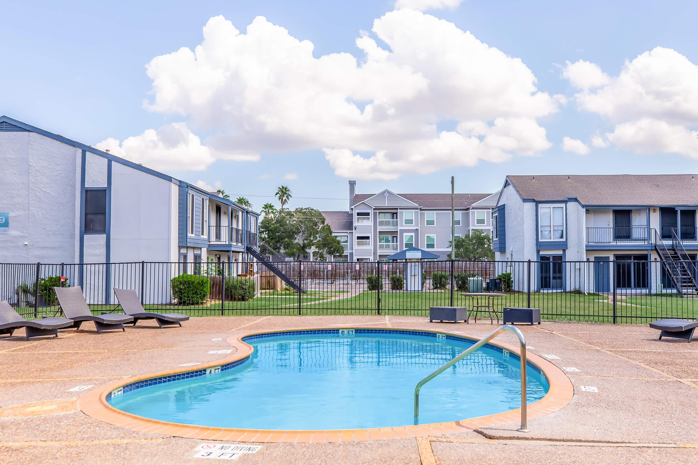 A clear blue swimming pool in a residential area, surrounded by lounge chairs. An apartment complex is visible in the background, with green grass and trees. The sky is partly cloudy, creating a bright and inviting atmosphere for relaxation and leisure.