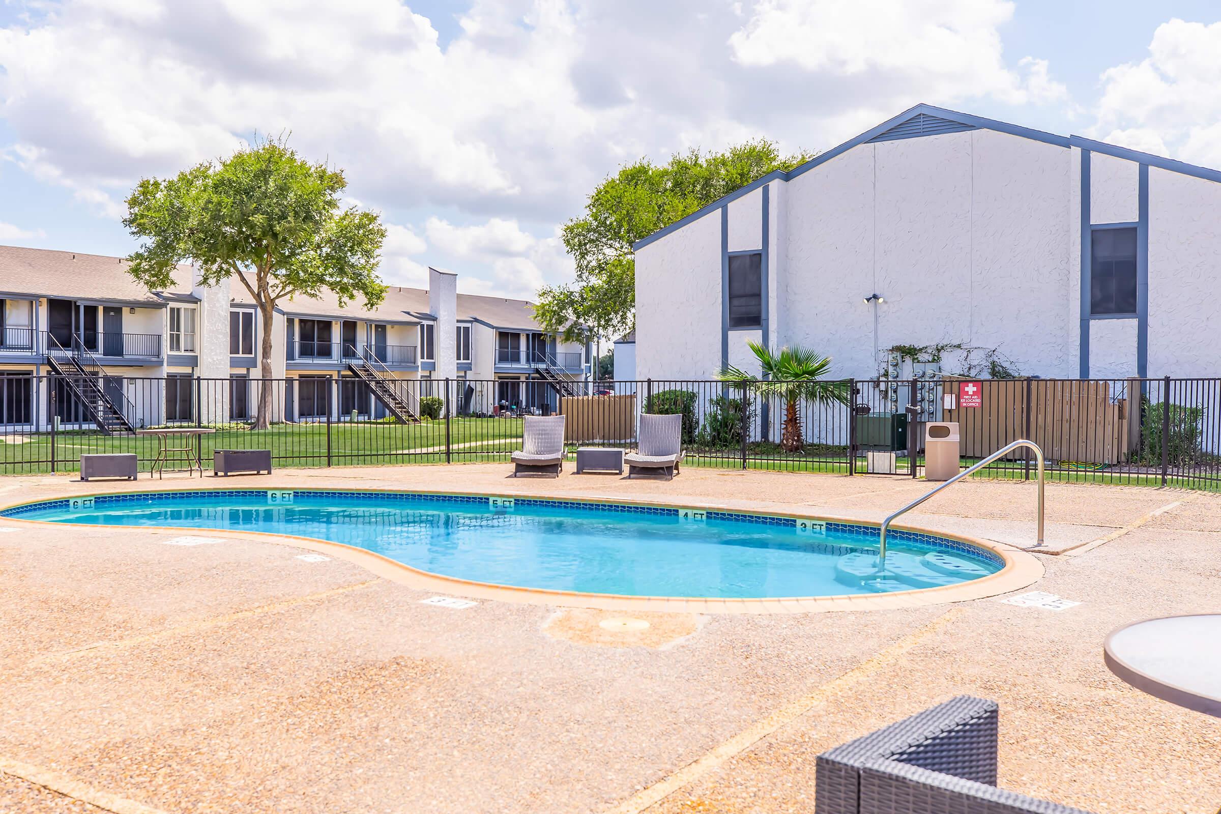 A clear swimming pool surrounded by lounge chairs, set in an outdoor area with green lawn and trees. In the background, two multi-story residential buildings can be seen under a partly cloudy sky. The pool area is enclosed by a fence, offering a serene and inviting atmosphere.
