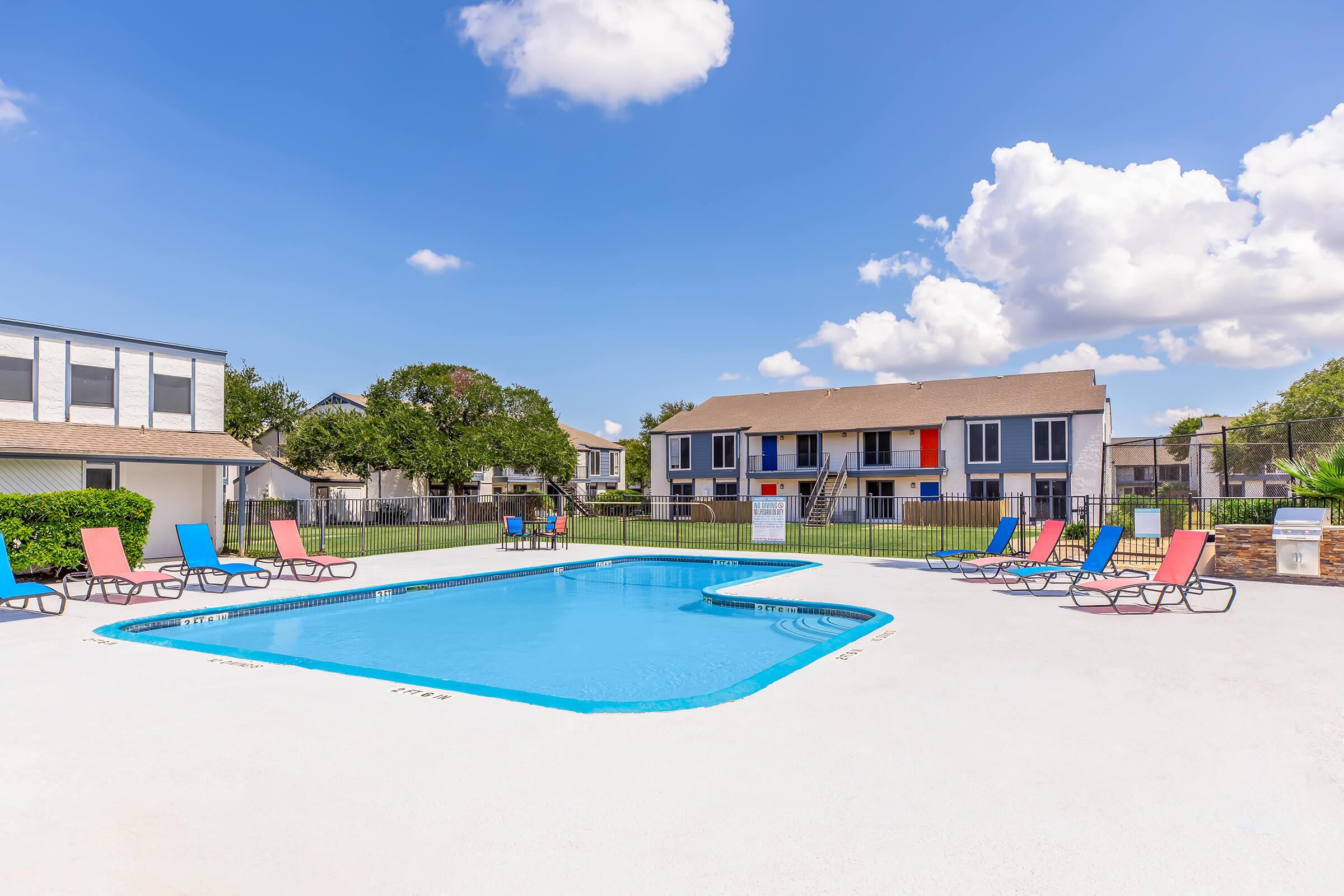 A bright and inviting pool area featuring a clear blue swimming pool surrounded by colorful lounge chairs. Lush greenery and residential buildings are visible in the background under a sunny sky with a few fluffy clouds. A peaceful and relaxing atmosphere ideal for leisure activities.