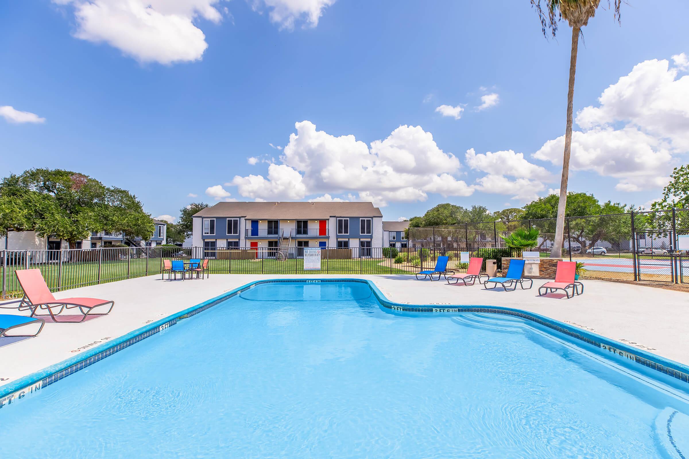A clear blue swimming pool surrounded by lounge chairs in a residential area. In the background, there are two-story apartment buildings with colorful balconies and a well-maintained lawn. The sky is bright with fluffy clouds, and a palm tree adds a tropical touch to the scene.