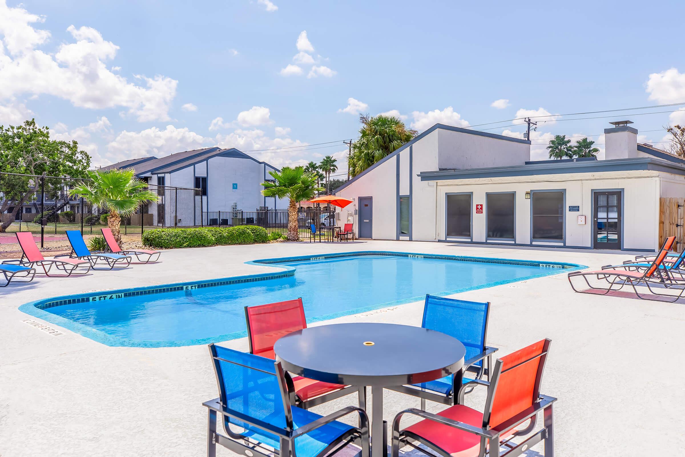 A bright, sunny pool area featuring a blue swimming pool surrounded by colorful lounge chairs in red, blue, and orange. Nearby is a shaded seating area with a table. The background includes residential buildings and palm trees, set against a clear blue sky with fluffy clouds.