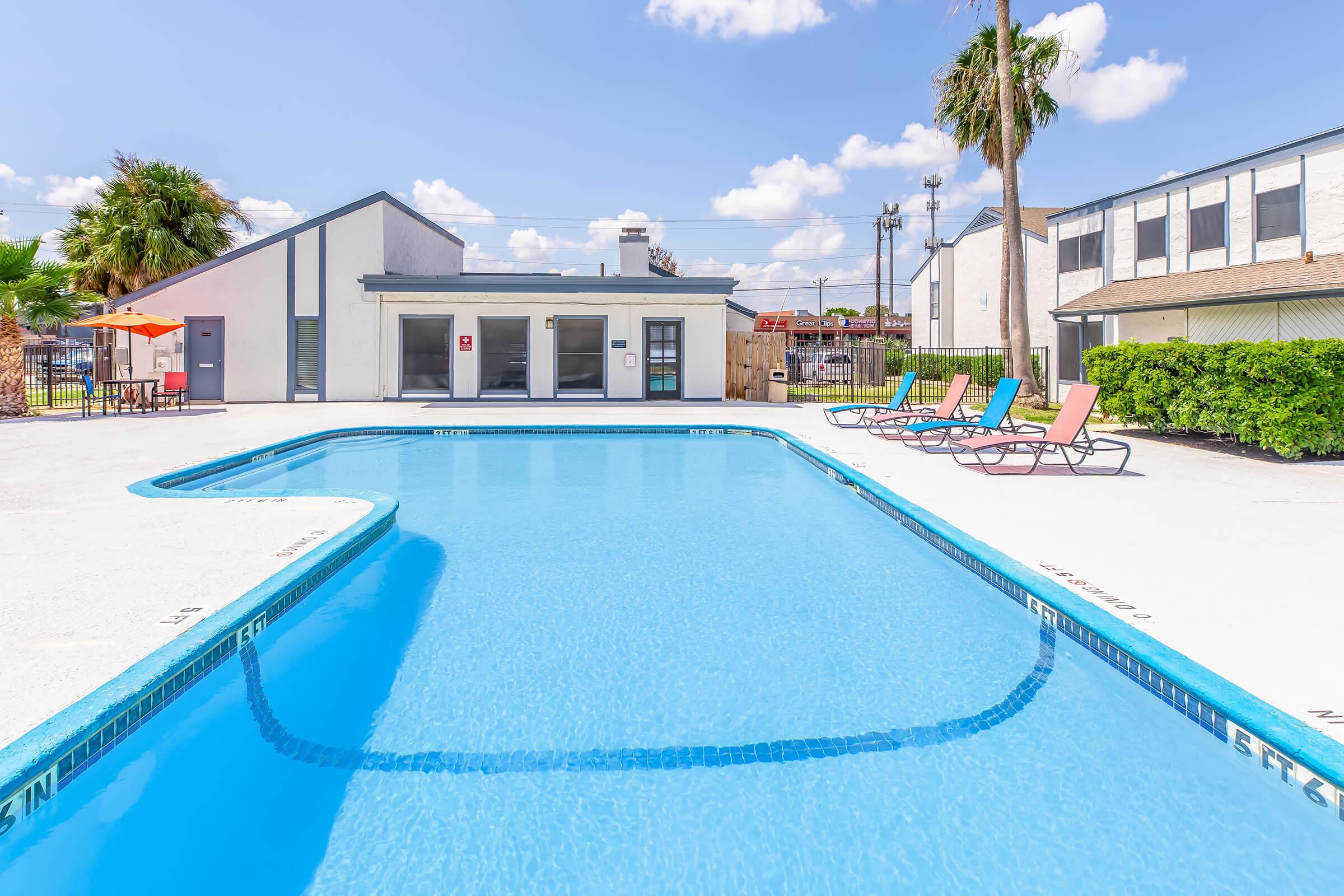 A sunny outdoor swimming pool surrounded by lounge chairs. Palm trees and a building are visible in the background, with a clear blue sky and fluffy white clouds above. The pool area is well-maintained, creating a relaxing atmosphere for residents.