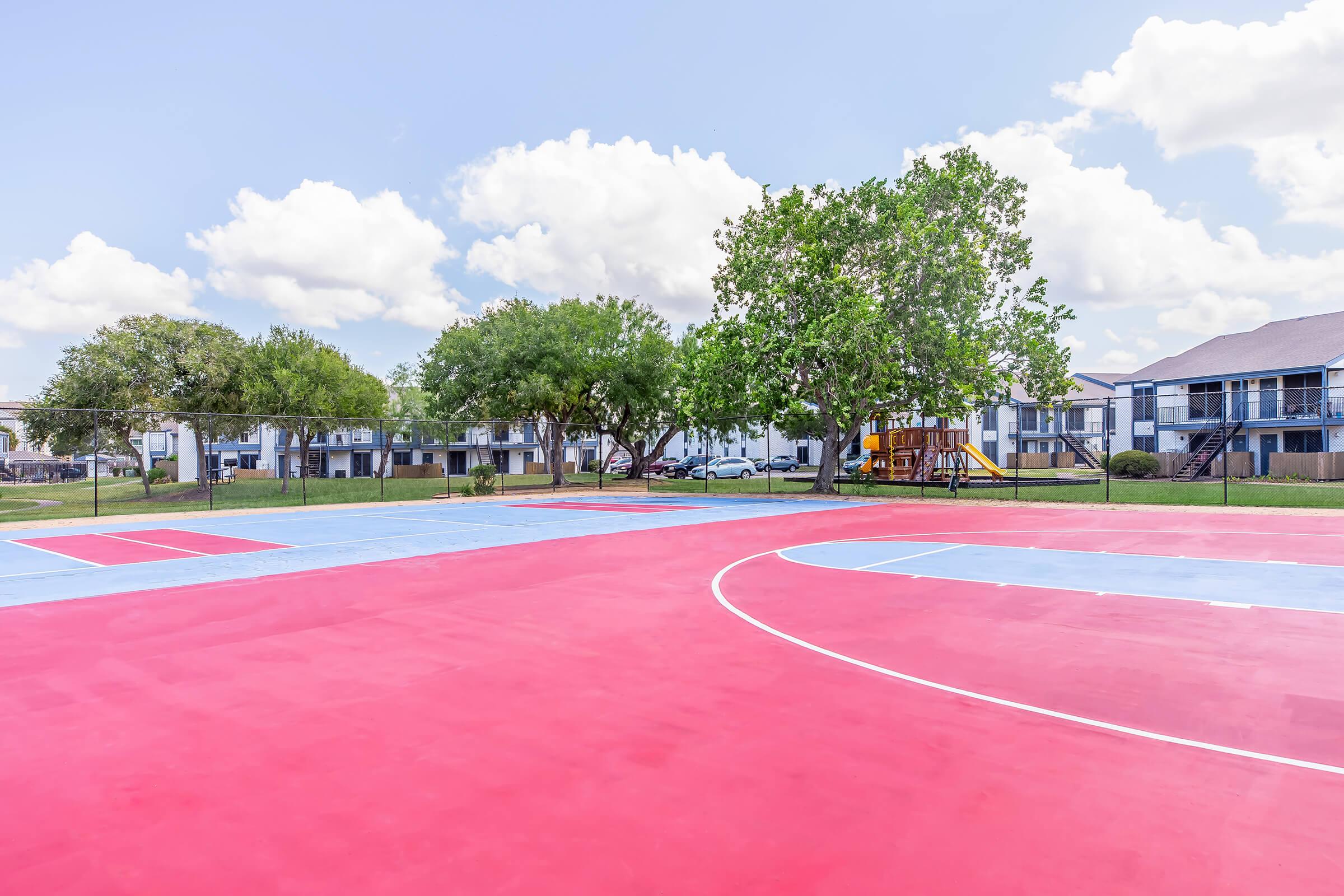 A wide view of an outdoor basketball court with red and blue sections, surrounded by grassy areas and trees. In the background, there are residential buildings and a playground visible. The sky is partly cloudy, creating a bright and sunny atmosphere.