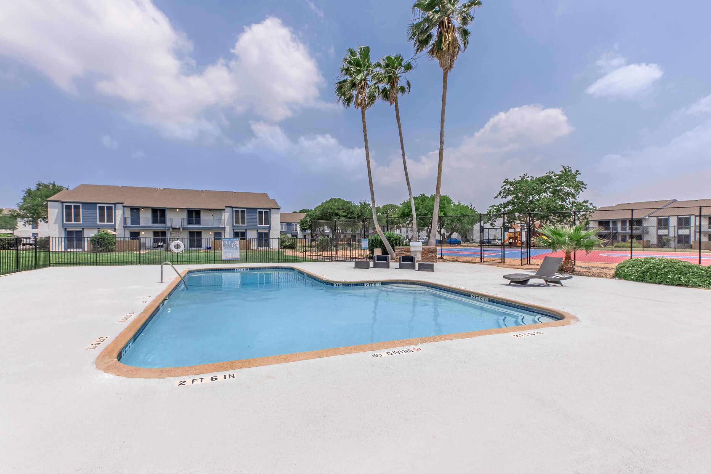 A serene outdoor swimming pool surrounded by palm trees and a fenced area, with lounge chairs nearby. In the background, there are multi-story apartment buildings and a playground, under a partly cloudy sky. The area is well-maintained and inviting for relaxation and leisure.