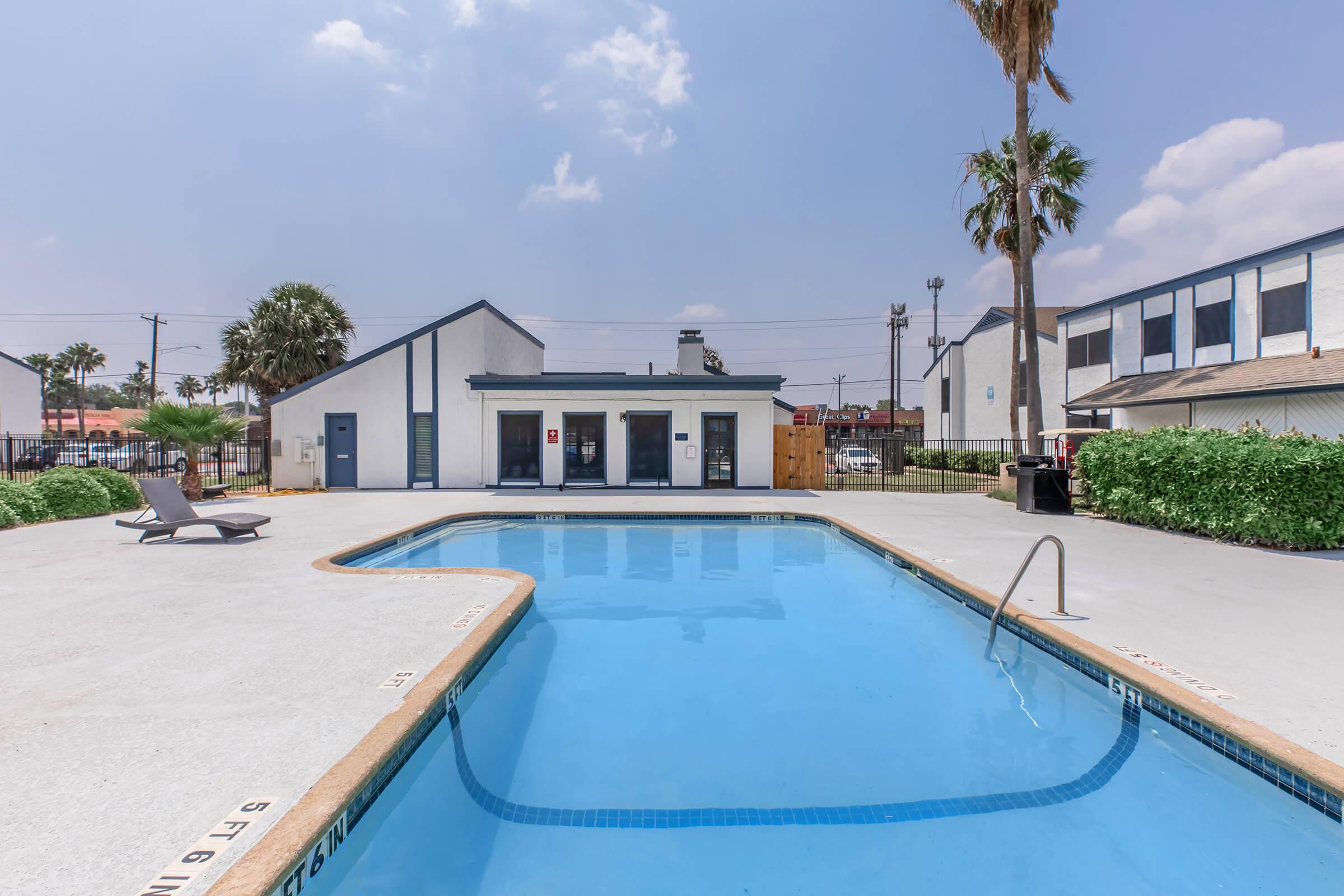 A clear pool surrounded by a concrete deck, with lounge chairs on one side. In the background, there's a building with multiple windows and palm trees. The sky is partly cloudy, adding a bright atmosphere to the scene.