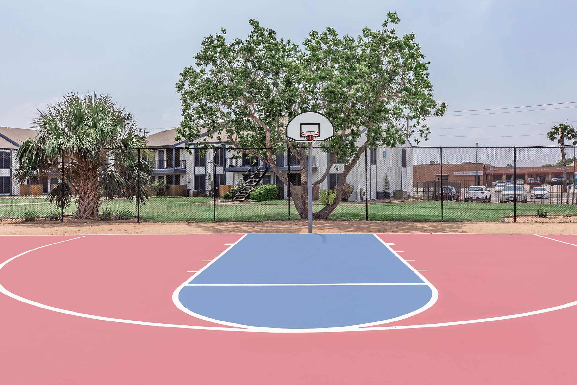 A basketball court with a blue half-court area and pink sidelines, surrounded by a chain-link fence. In the background, there is a large tree and residential buildings. The sky appears overcast, creating a calm atmosphere. The court is empty, emphasizing its openness and inviting nature.