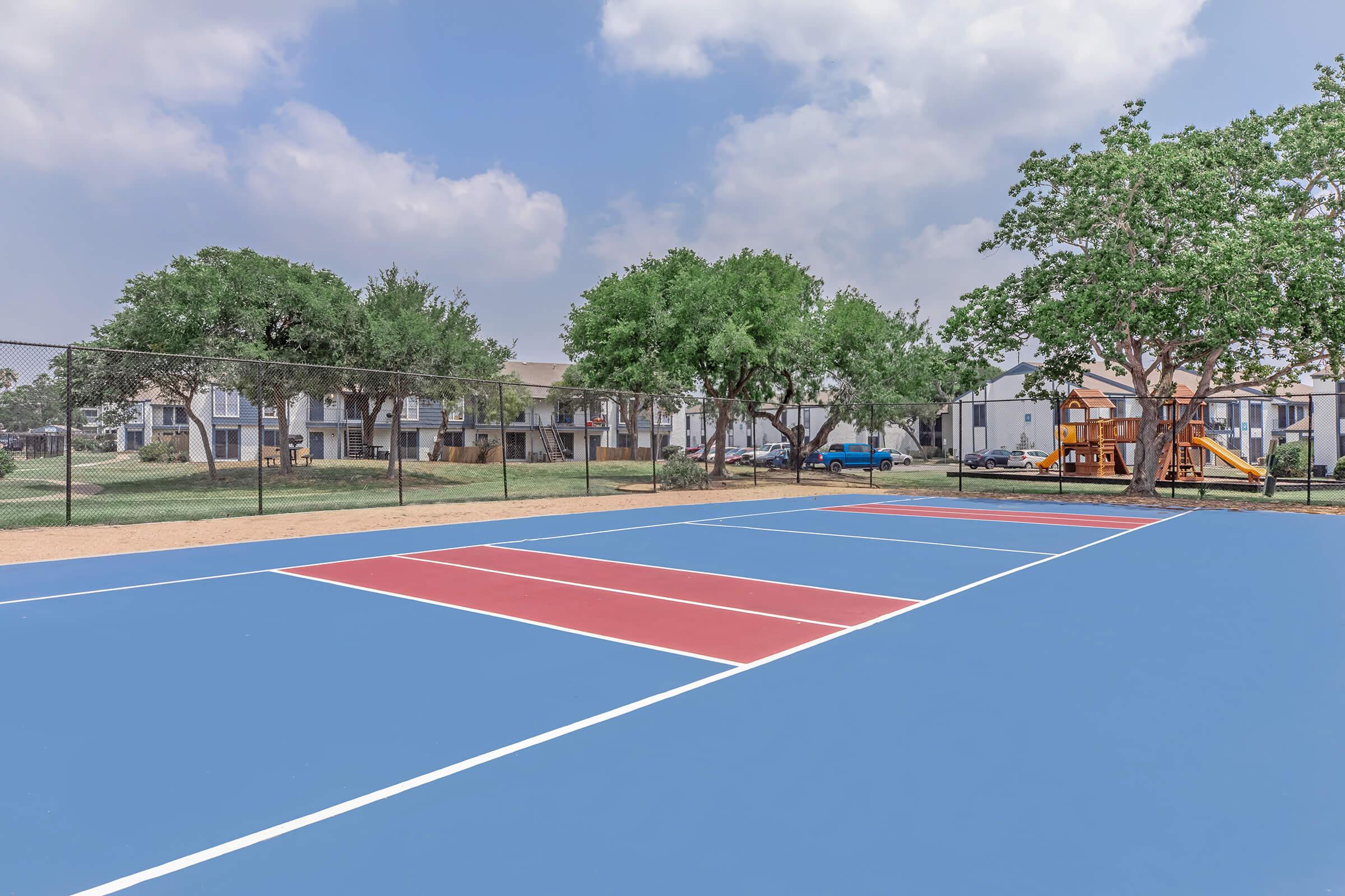 A brightly colored outdoor basketball court featuring blue, red, and white markings. In the background, there are several green trees and a playground structure, with residential buildings visible behind the court under a partly cloudy sky.