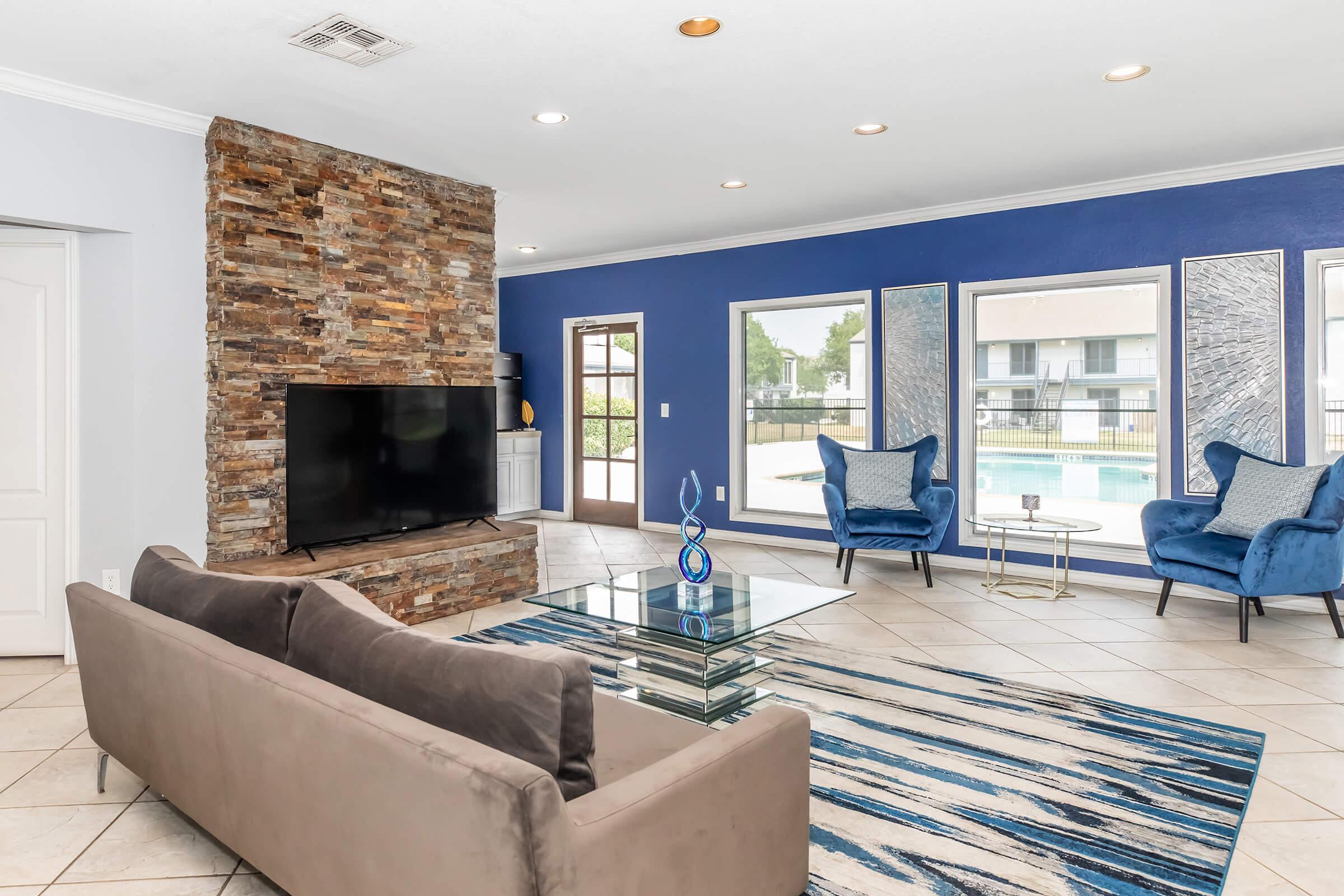 A modern living room featuring a stone wall, a large flat-screen TV, and a glass coffee table. The space includes a beige sofa and two blue accent chairs, with a patterned area rug on the tiled floor. Large windows provide views of an outdoor pool area, while a vibrant blue wall adds a pop of color.