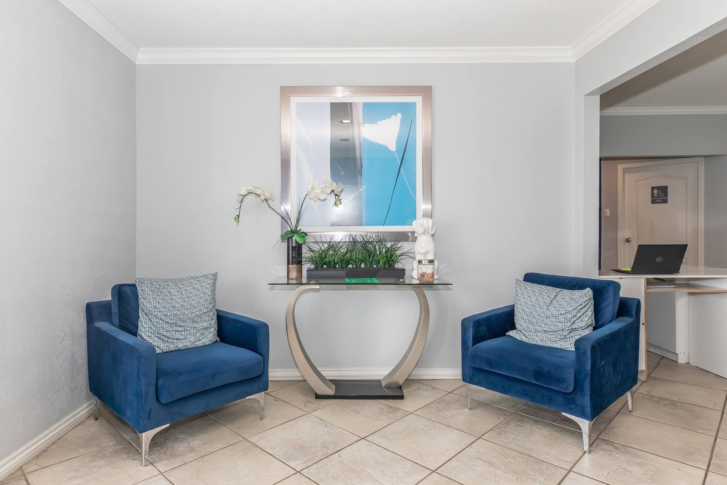 Two blue upholstered armchairs are positioned on either side of a modern glass console table. The table has a small green plant and a decorative statue. Above it hangs a framed mirror reflecting the room. The walls are light gray, and the floor features beige tiles. A doorway is visible in the background.