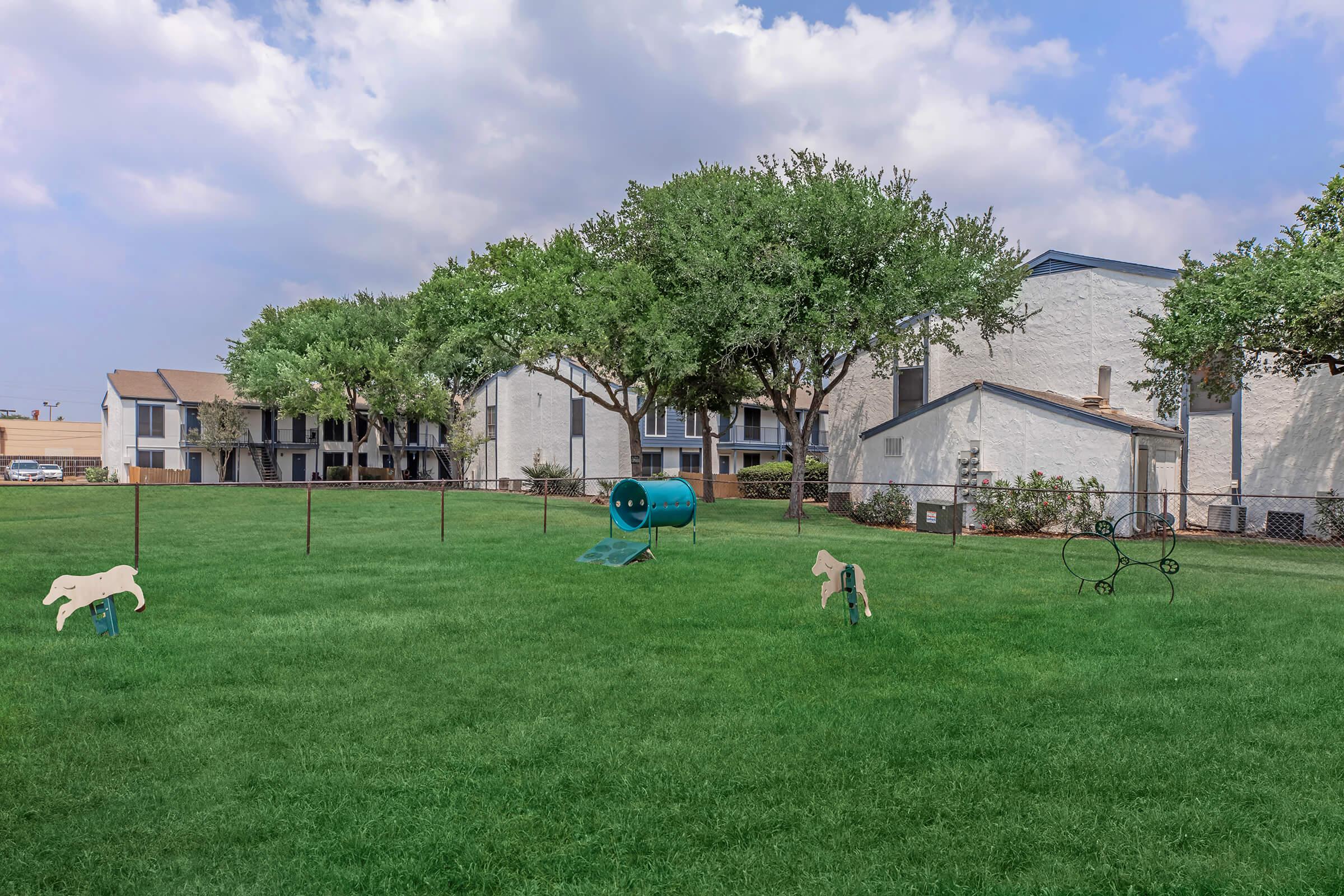 A grassy area of a dog park featuring agility equipment such as a tunnel and obstacles, surrounded by leafy trees and residential buildings in the background under a partly cloudy sky.