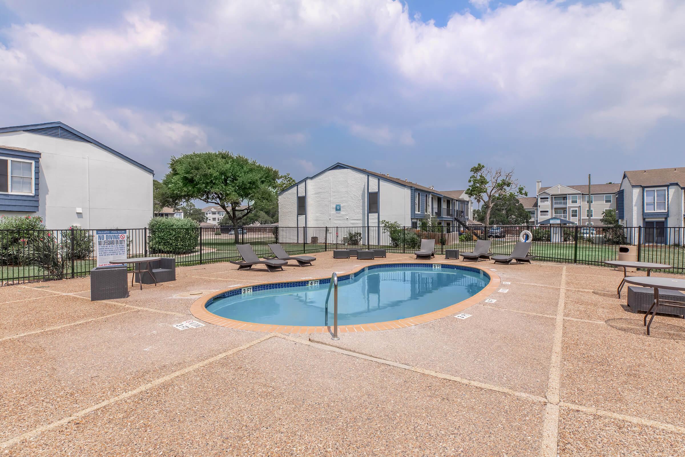 A residential swimming pool area featuring an oval-shaped pool surrounded by lounge chairs. The pool is enclosed by a fence, and there are apartment buildings in the background under a partly cloudy sky. The setting is well-maintained with greenery around the pool area.