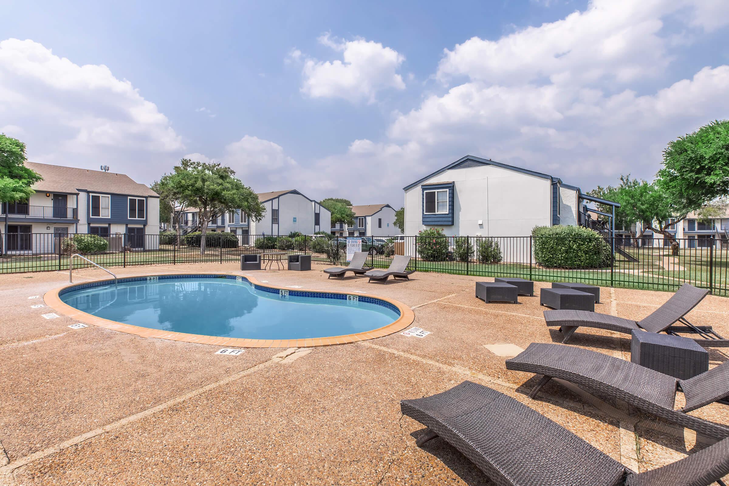 A swimming pool surrounded by lounge chairs, with residential buildings in the background. The pool area is enclosed by fencing, and there are green trees and shrubs nearby. The sky is partly cloudy, creating a bright and inviting atmosphere.