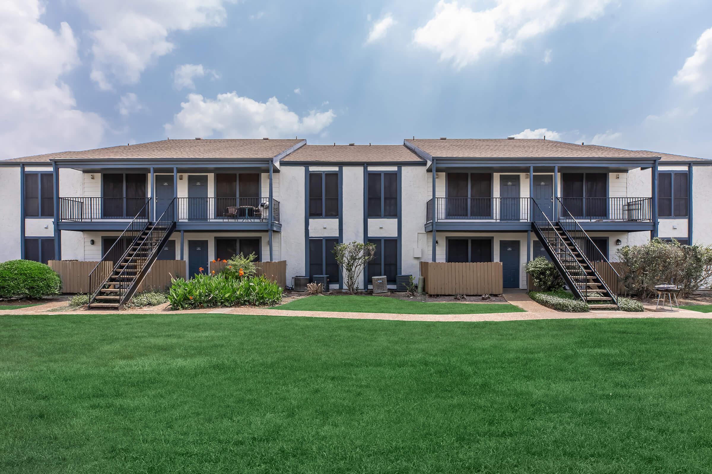 Two-story apartment buildings with blue accents, featuring balconies and staircases. The foreground has well-manicured green grass and flower beds, while the background shows a cloudy sky. The buildings are symmetrical and have a welcoming appearance, set in a landscaped environment.