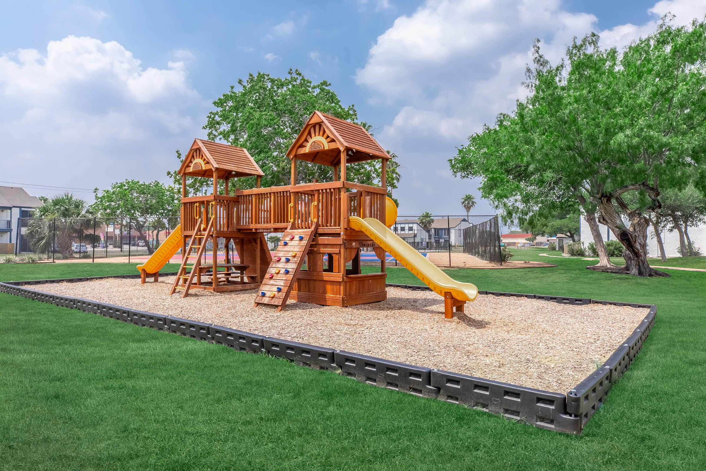 A wooden playground structure with two raised playhouses, featuring slides and climbing areas, surrounded by green grass and trees. The sky is partly cloudy, creating a bright and inviting outdoor scene.