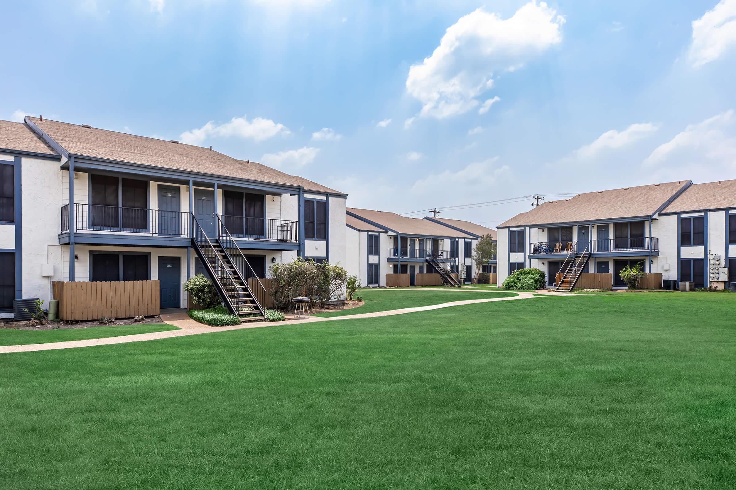 A well-maintained apartment complex featuring two-story buildings with balconies, surrounded by a lush green lawn. Clear blue sky with scattered clouds can be seen in the background, creating a bright and inviting atmosphere. Pathways connect the buildings, enhancing the outdoor space.