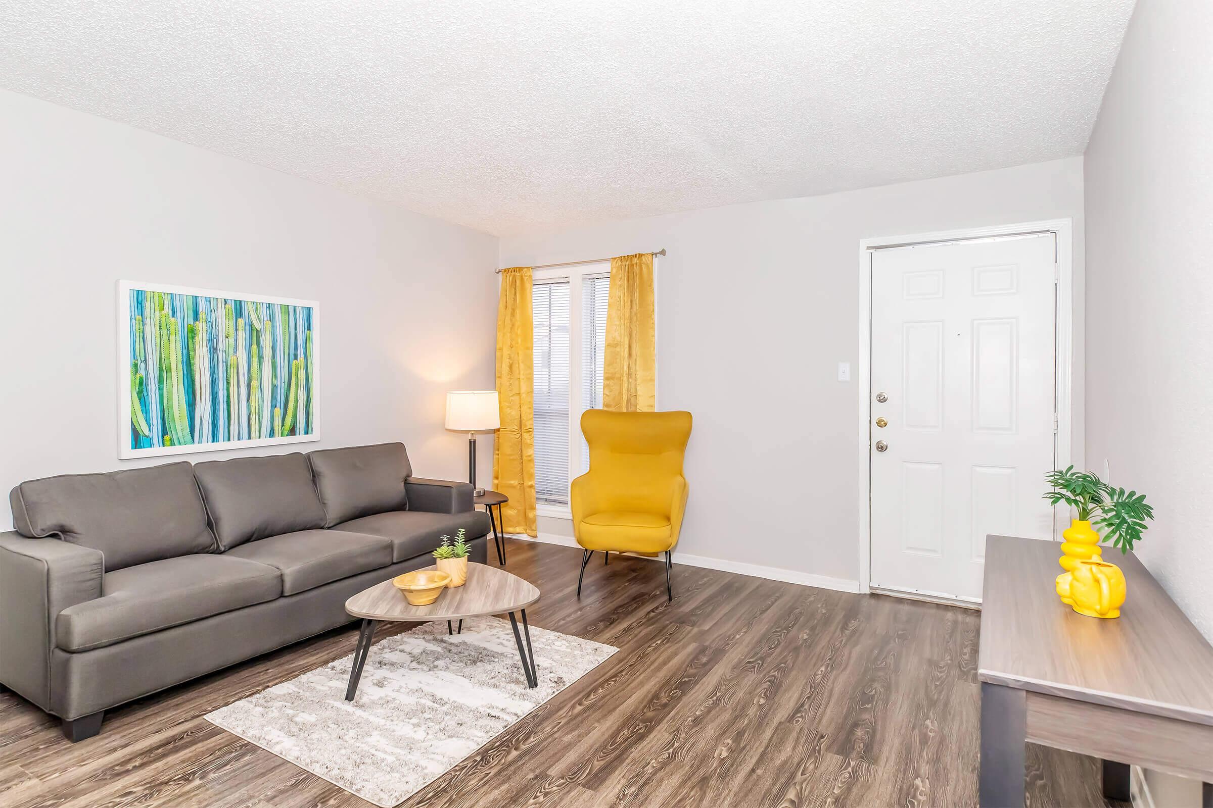 A cozy living room featuring a gray sofa, a yellow accent chair, a round coffee table, and a small plant. A colorful artwork with vertical lines hangs on the wall, complemented by sheer curtains. A light fixture stands beside the sofa, and a console table with a decorative object sits against the wall near the door.