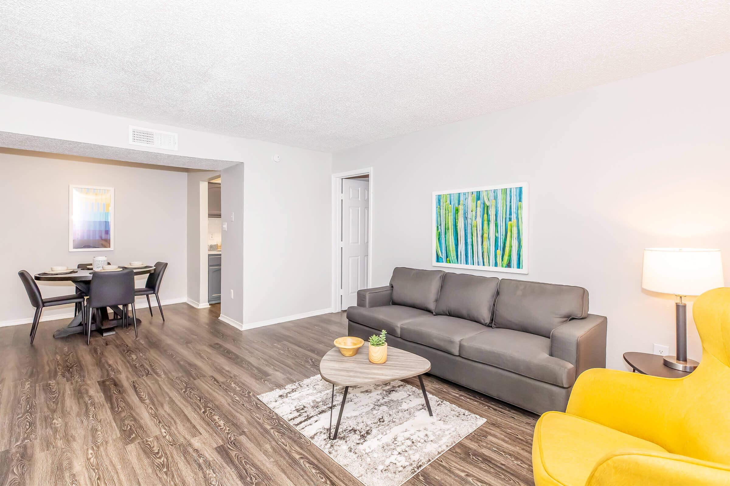 A modern living room featuring a gray sofa, a round coffee table with a small plant, and a yellow accent chair. A colorful abstract painting of vertical lines is on the wall, and a dining area with a table and chairs is visible in the background. The space has a neutral color scheme and wooden flooring.