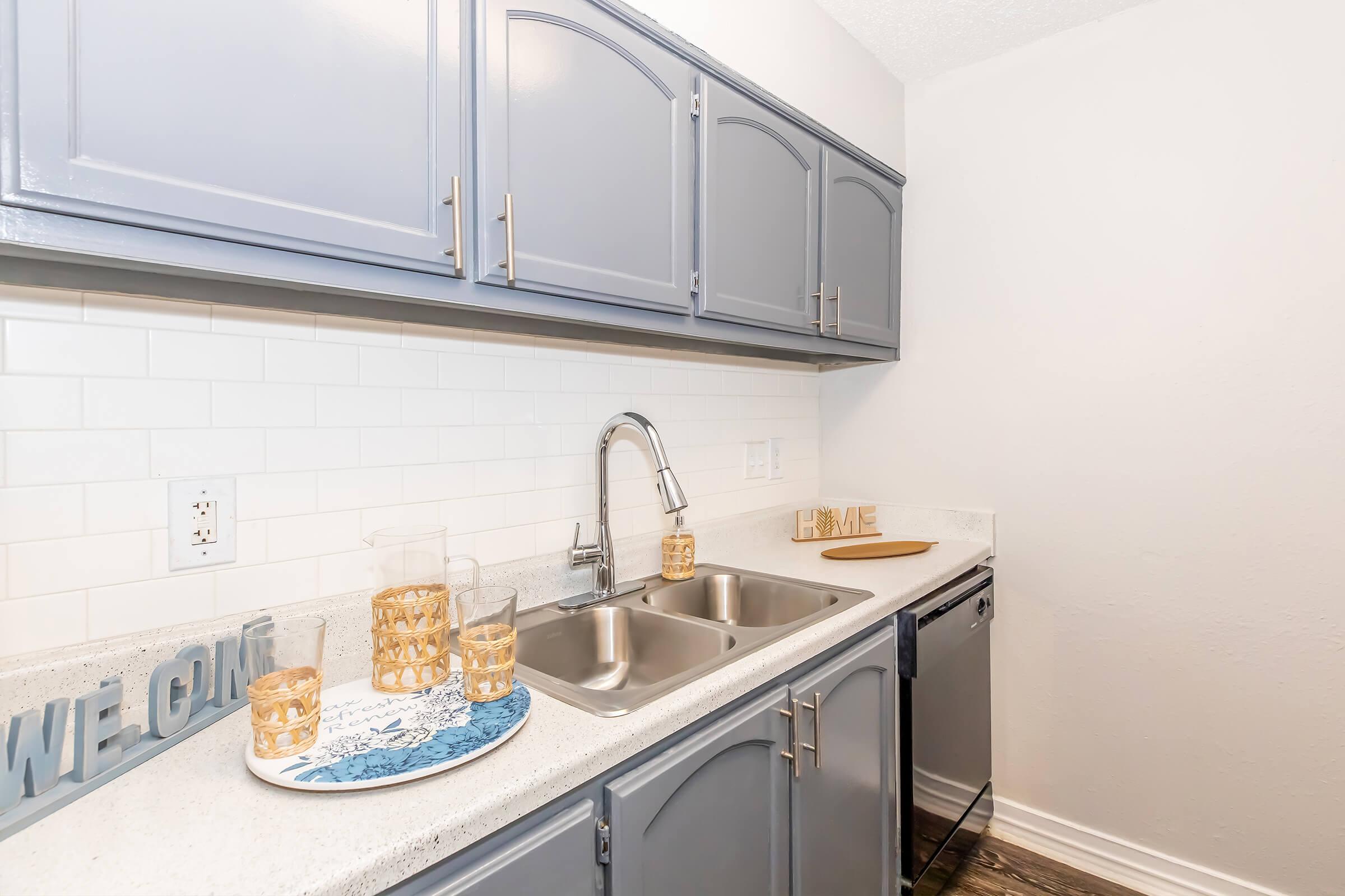 A modern kitchen featuring gray cabinets, a white subway tile backsplash, and a stainless steel sink. On the countertop, there are decorative items including a blue and white plate, golden glass holders, and a wooden cutting board. The overall aesthetic is clean and inviting.